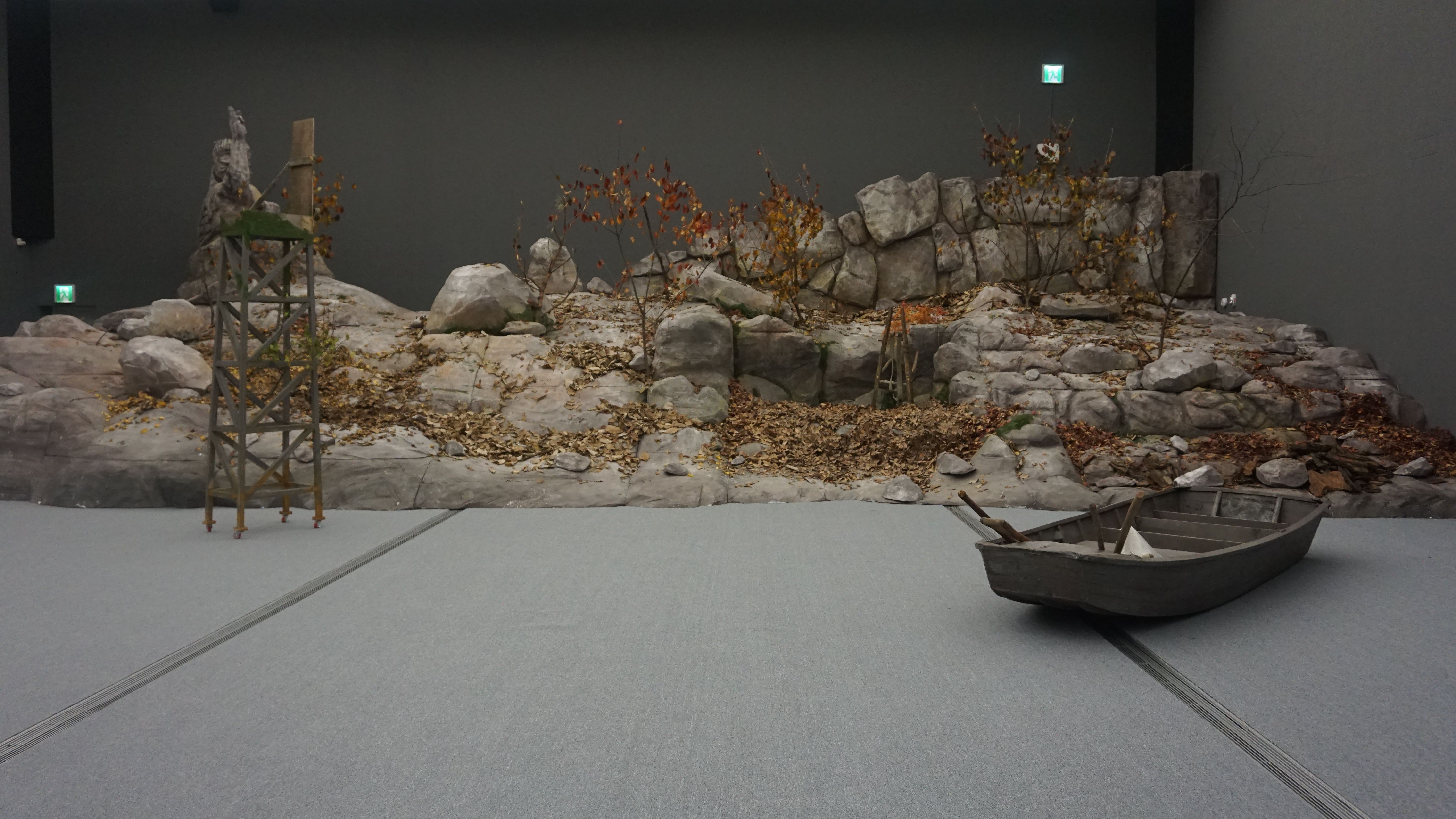 An art installation featuring a rugged rock shore with a wooden boat positioned on the floor, mimicking a natural shorelin...