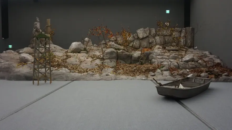 An art installation featuring a rugged rock shore with a wooden boat positioned on the floor, mimicking a natural shorelin...