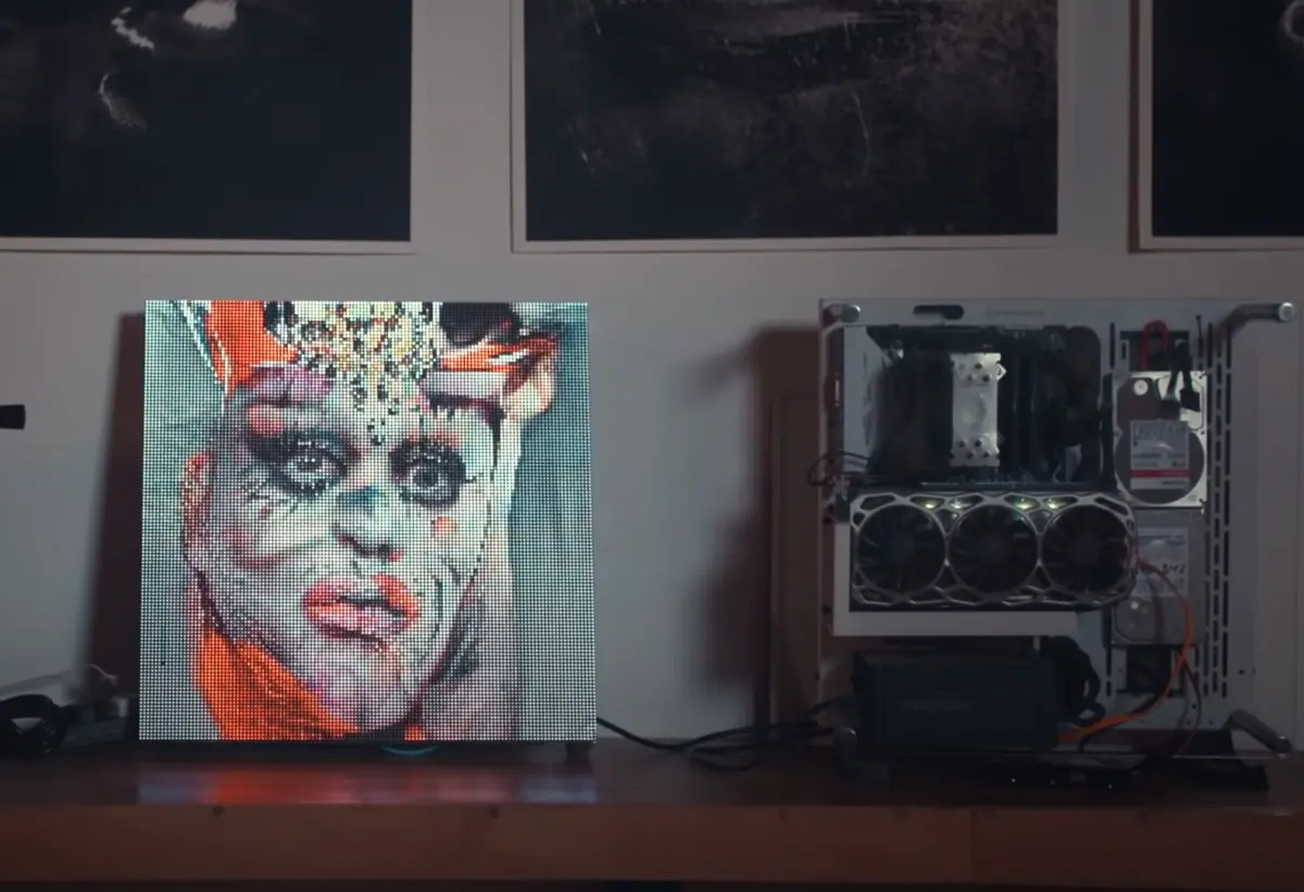 A colorful painted canvas of a figure adorned with make-up and ornaments is displayed in an interior setting. An vintage recording device stands near the artwork and three black and white framed posters partially peek from the top of the image frame.