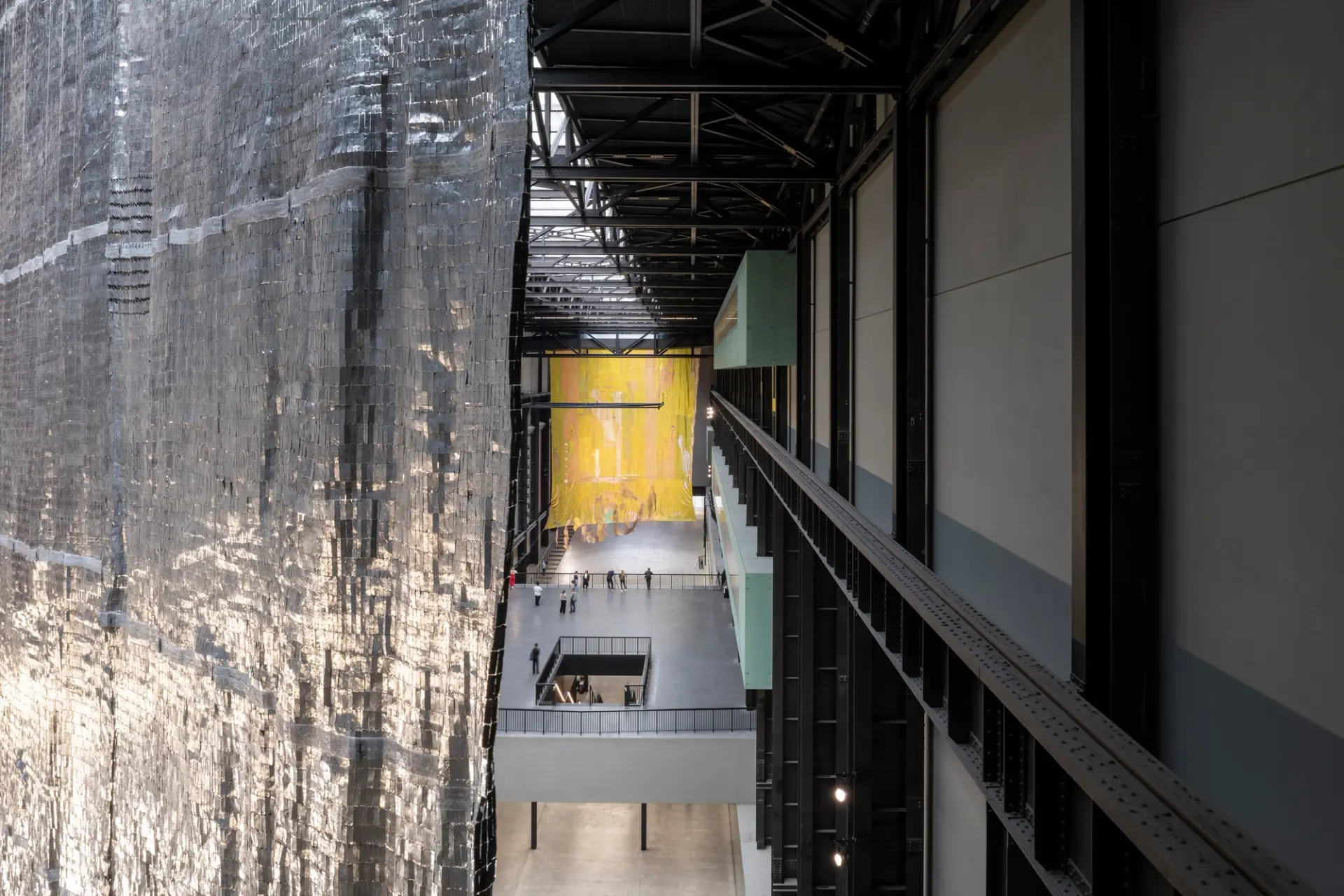 Aerial view inside a modern gallery space featuring a massive shimmering silver installation on the left and a large yellow art piece in the distance, with visitors walking below.