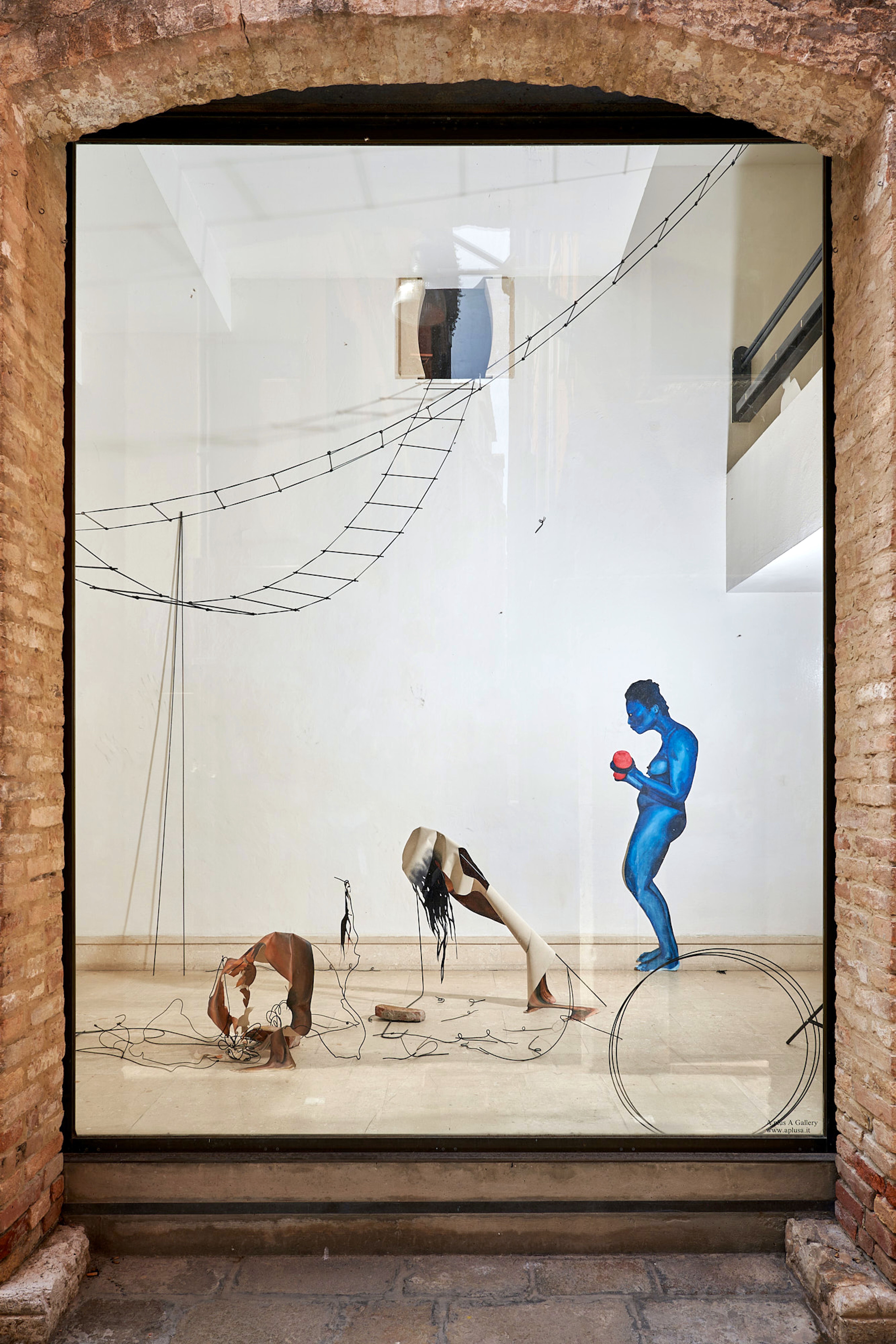 A window display of an art installation with wire ladders and abstract human forms, including a blue-painted figure holding a red object.