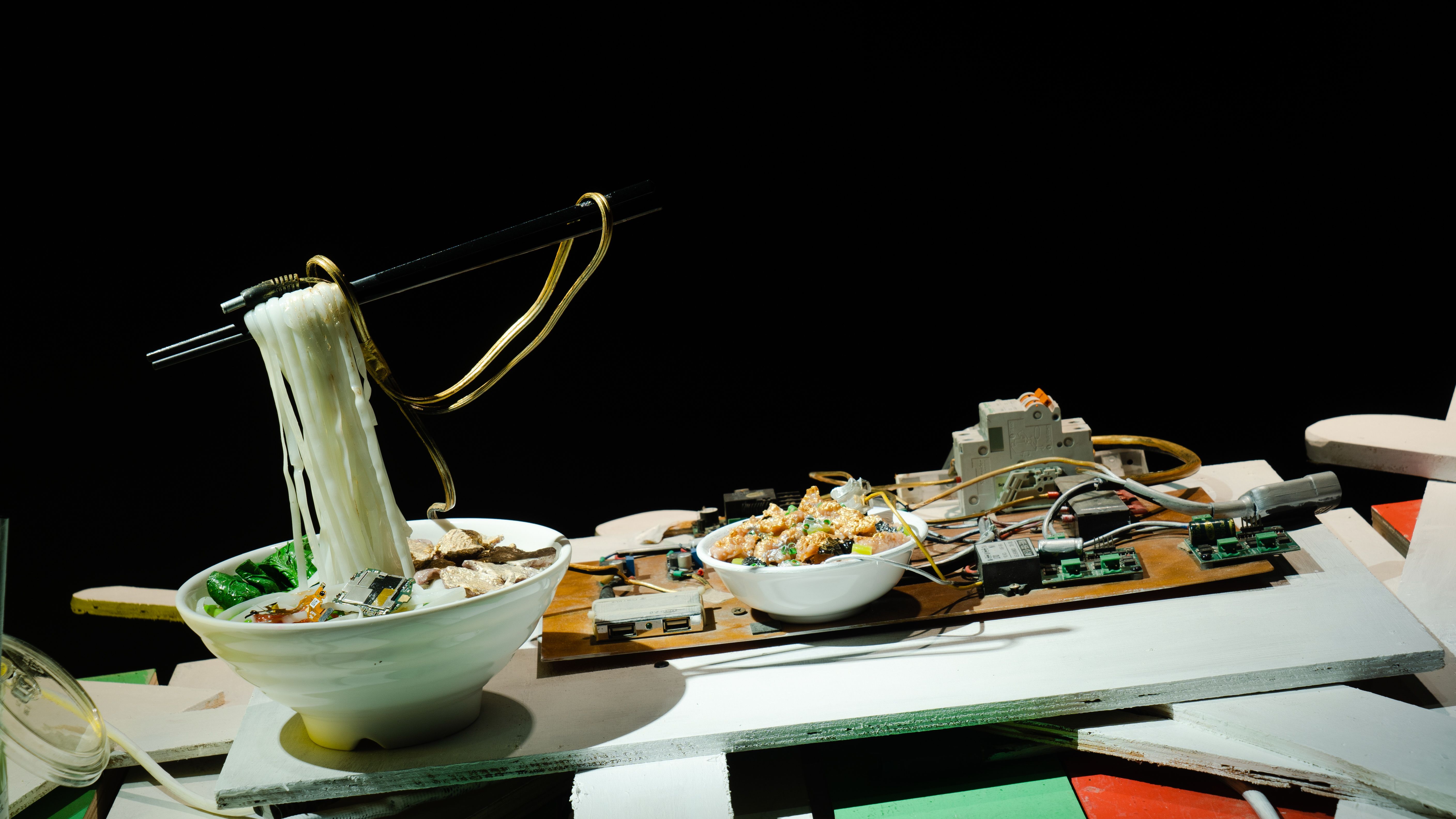 Robotic arms serving noodles in a bowl on a messy table with electronic parts.