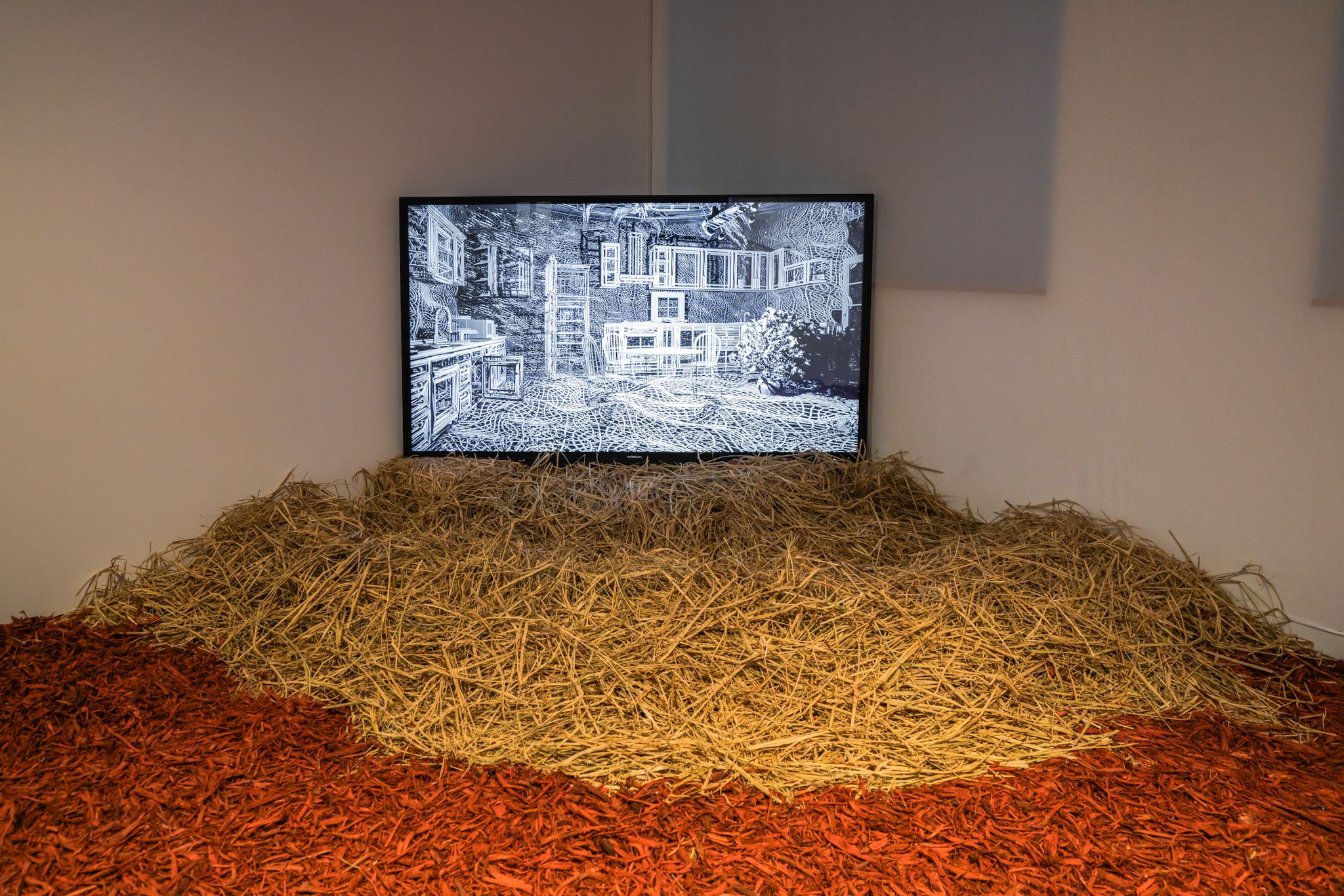 An installation featuring a TV screen displaying a black and white drawing. The TV is surrounded by hay and red mulch.