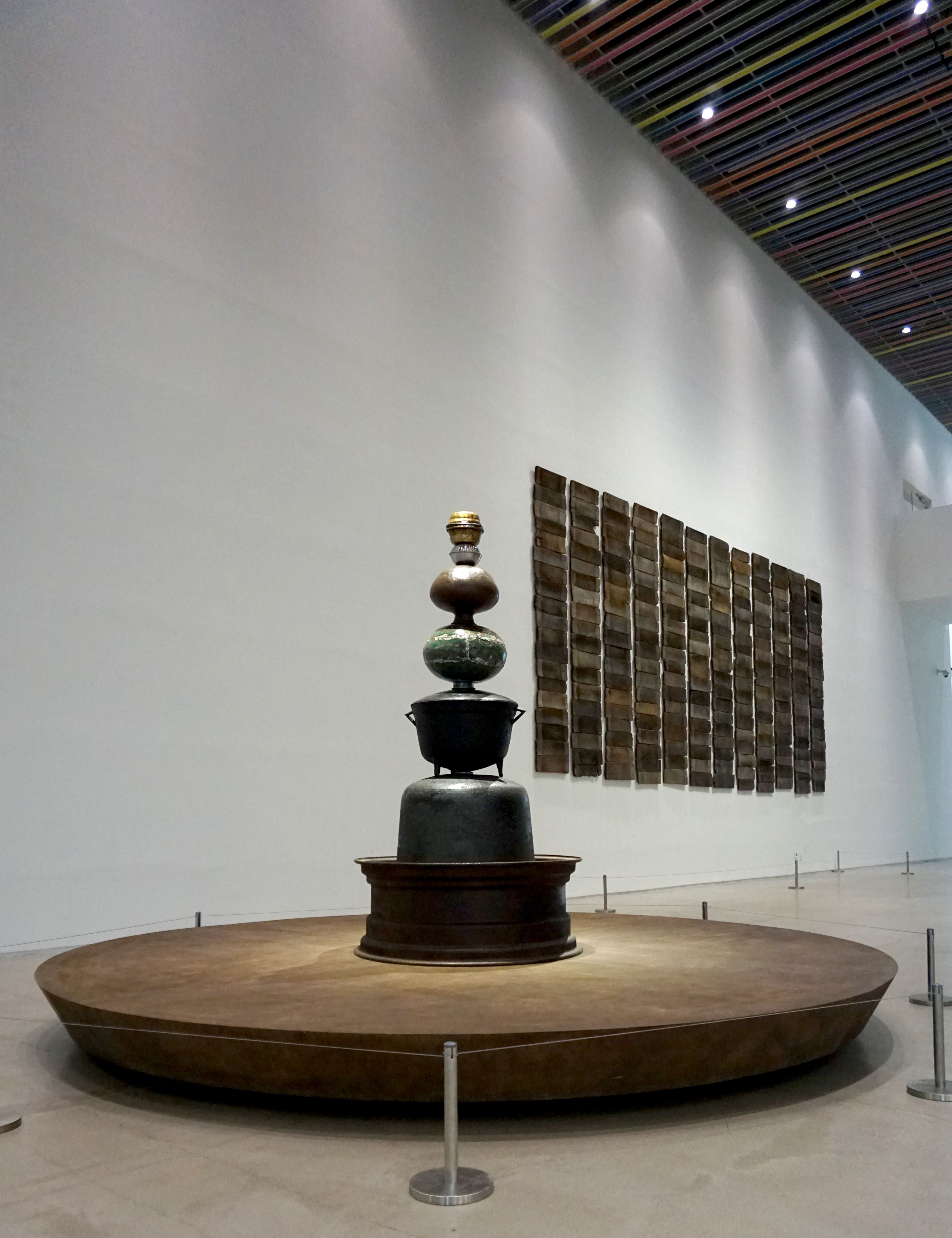A spacious gallery displays a large mixed-media sculpture in the center of the room, with accompanying wall art in the sur...