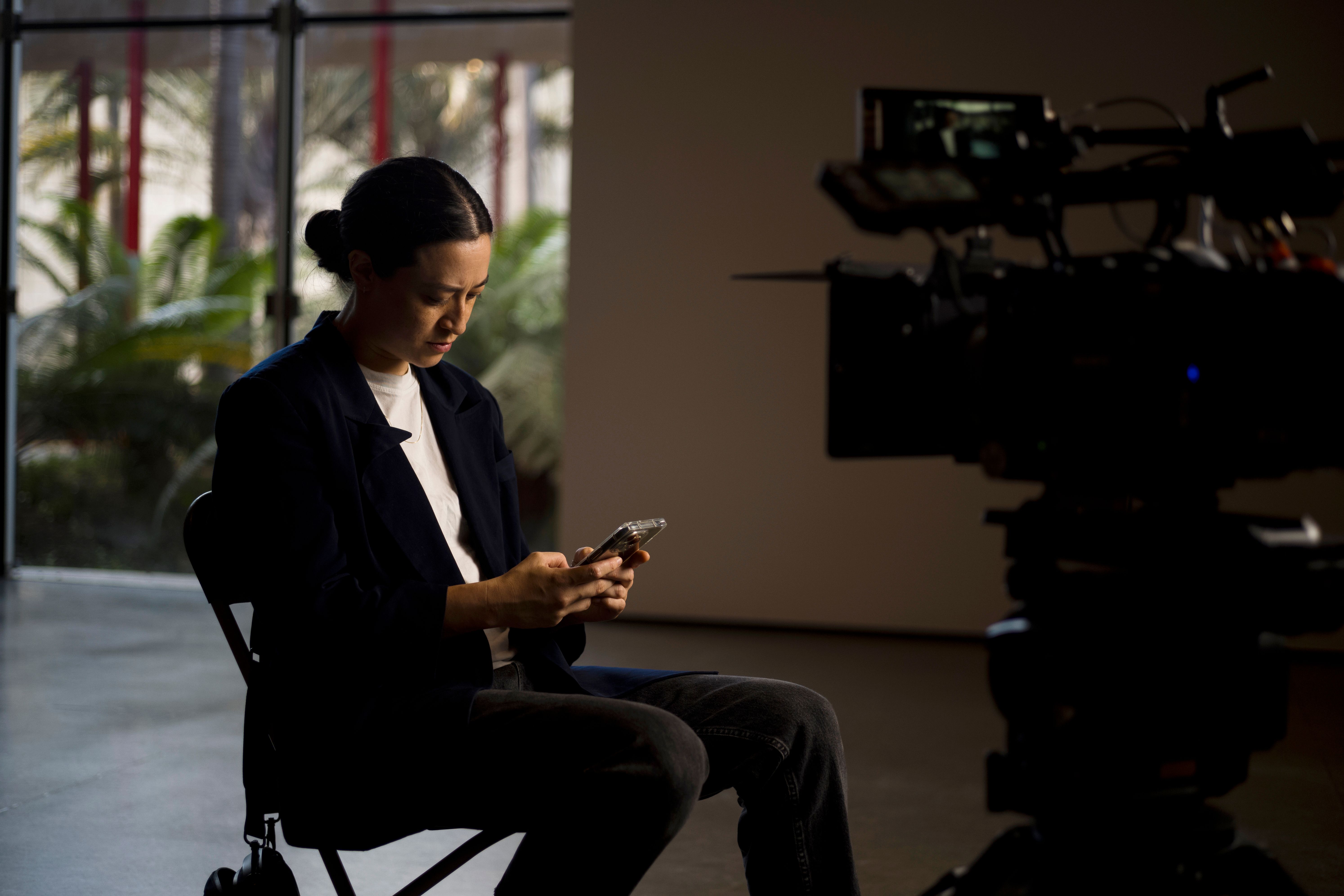 A person sits on a chair indoors, looking down at their phone, with a large camera set up nearby.