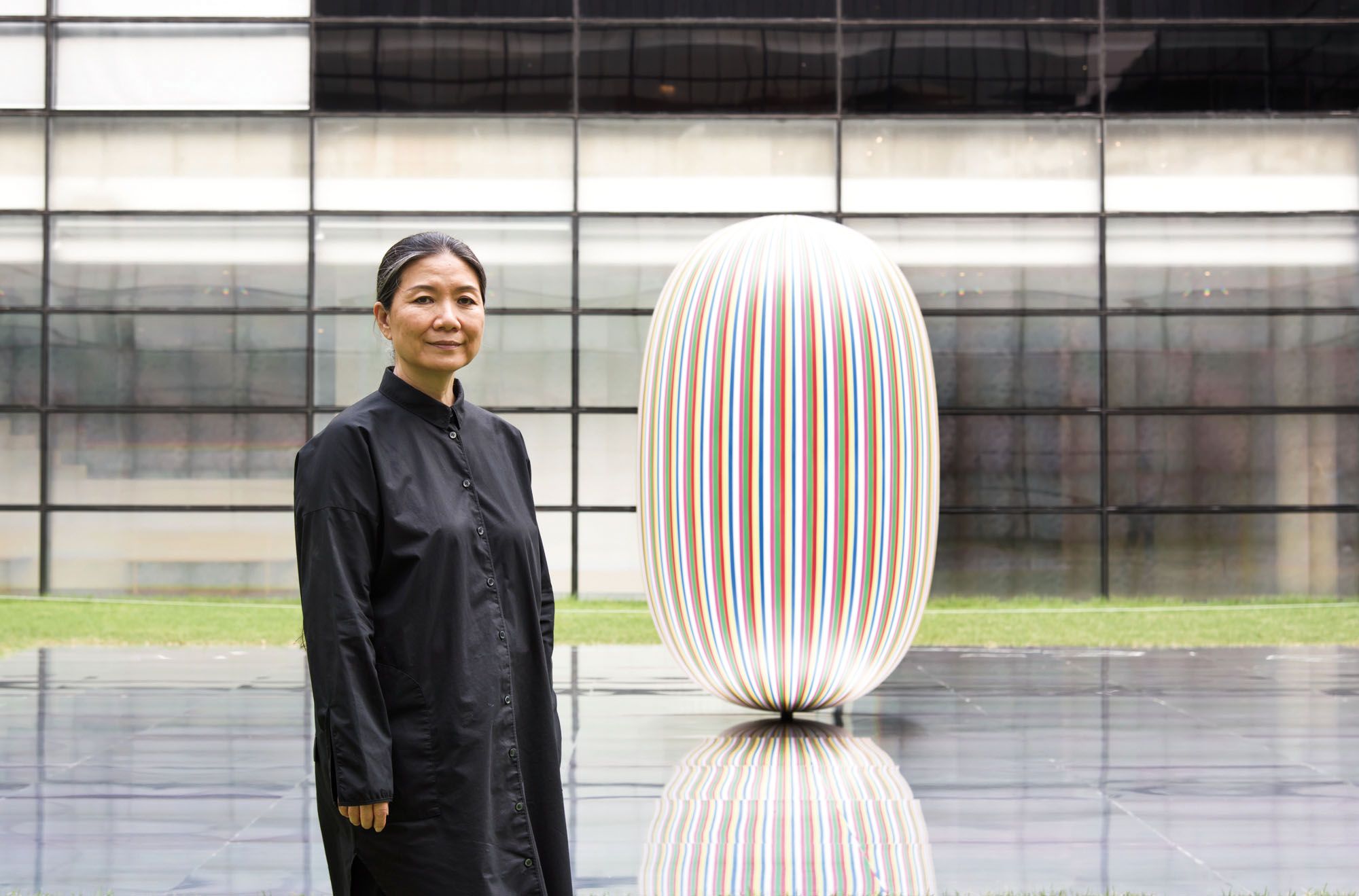 A person dressed in black stands before a glossy elliptical sculpture situated on a mirrored rectangular platform. The sce...