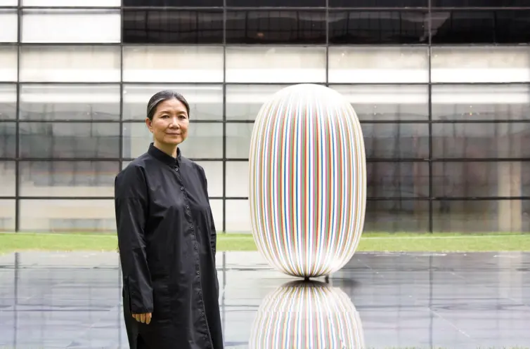 A person dressed in black stands before a glossy elliptical sculpture situated on a mirrored rectangular platform. The sce...