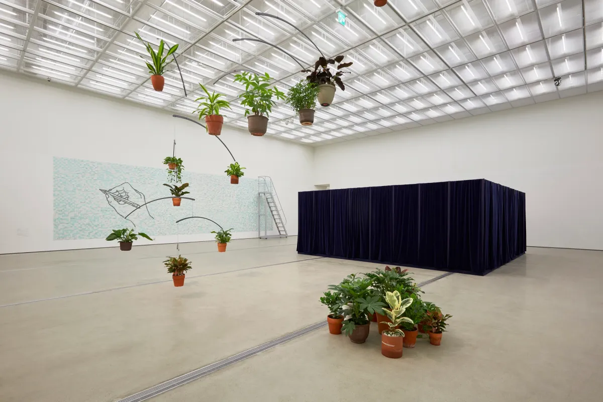 Three distinctive art installations in a brightly lit gallery: hanging pots with houseplants intertwined with bent wooden sticks, a wide rectangular aqua blue piece featuring a sketch of a hand holding a pencil, and a large, metallic ladder leaning against a dark blue cubic structure shrouded in curtains.