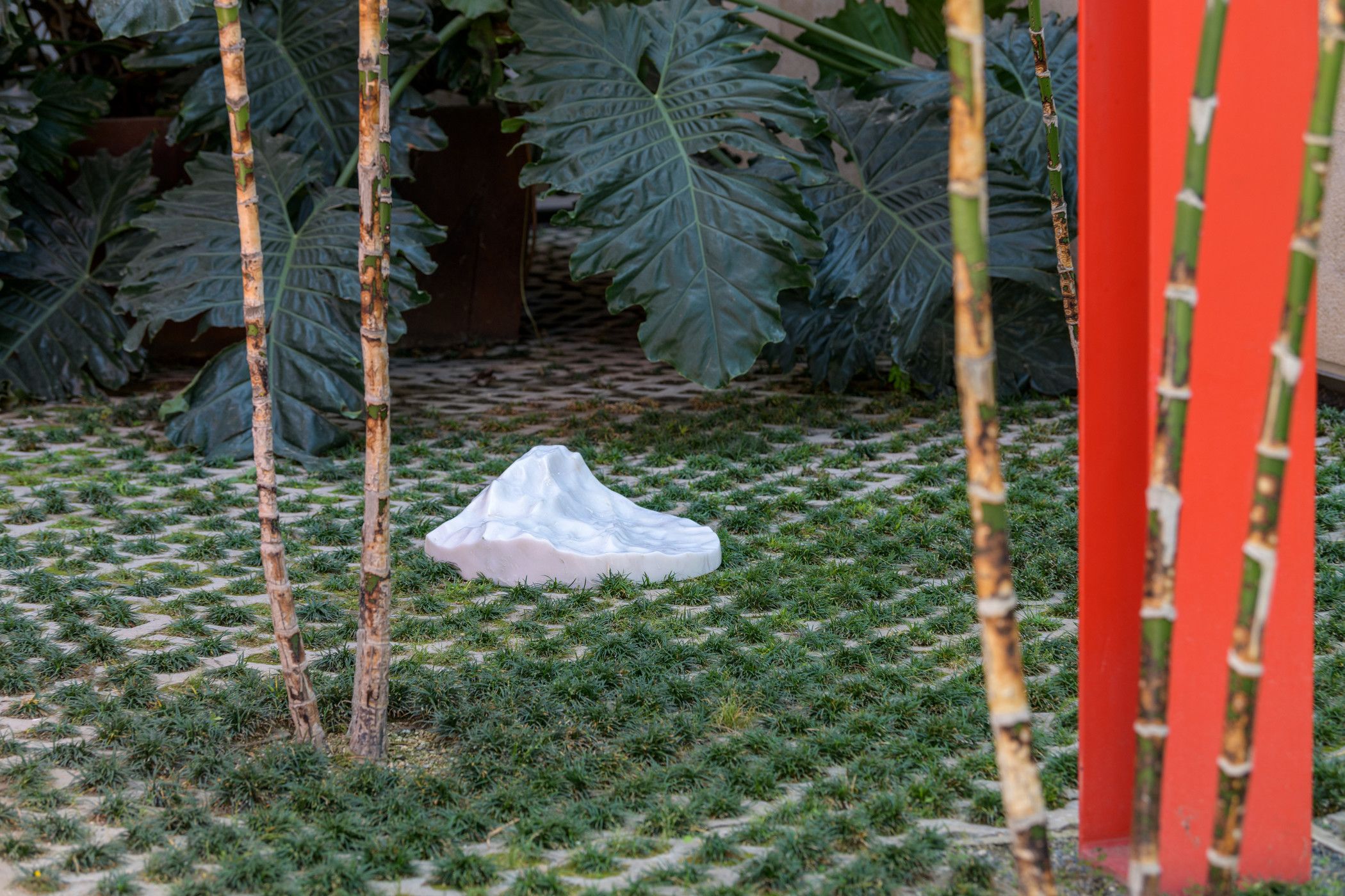 A white, amorphous sculpture surrounded by landscaping greenery and trees. Red stick forms protrude from the ground around...