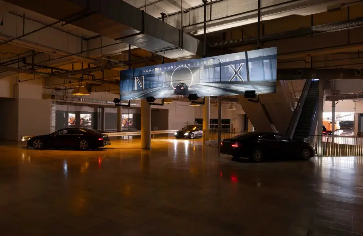 Luxury cars are displayed in a dim, industrial exhibition space beneath a large illuminated banner reading “Home of Nonhuman Excellence.”