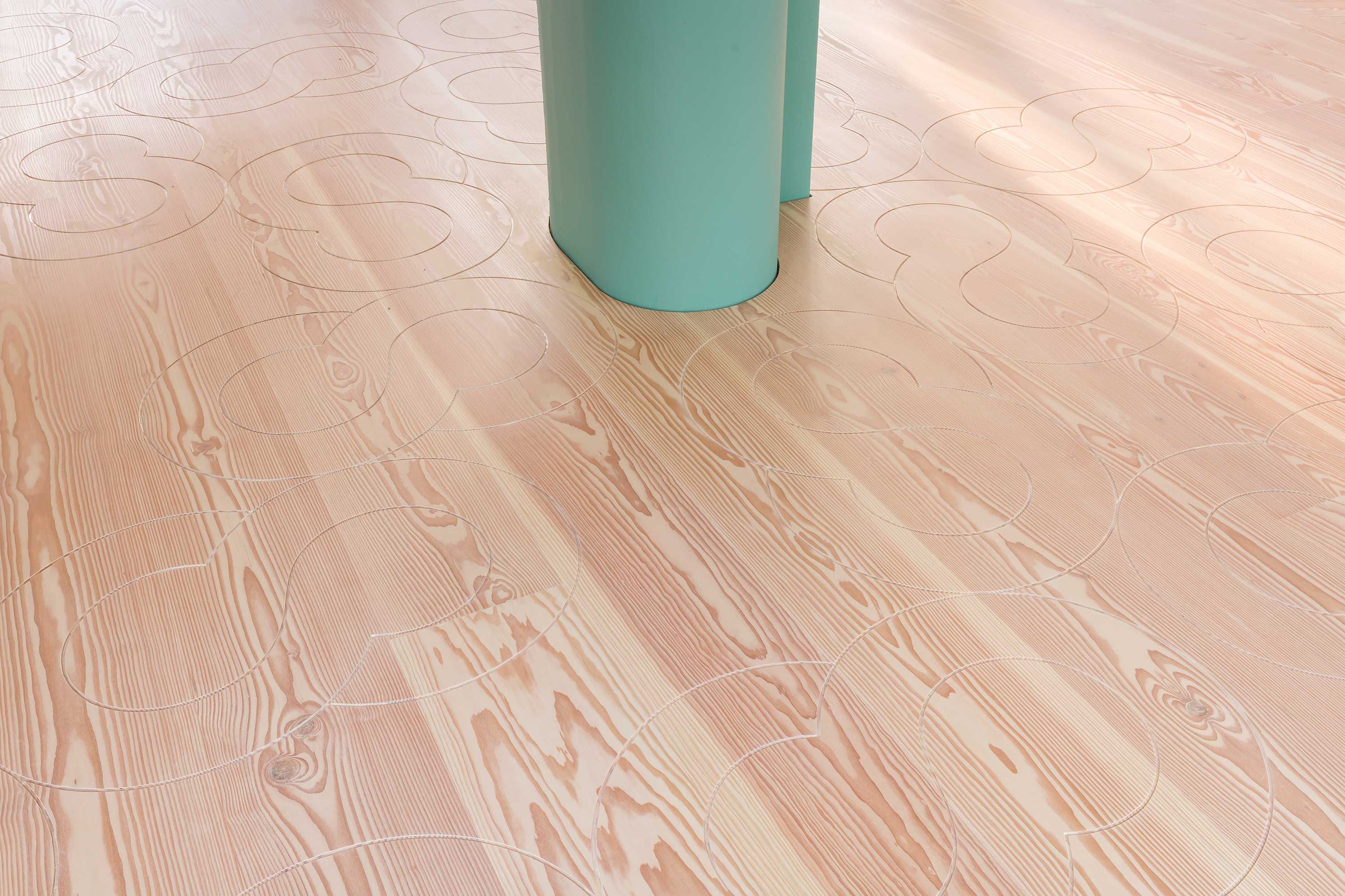 Detail of a light wooden floor with engraved circles and a pastel teal column in the corner, complementing the wooden tones.
