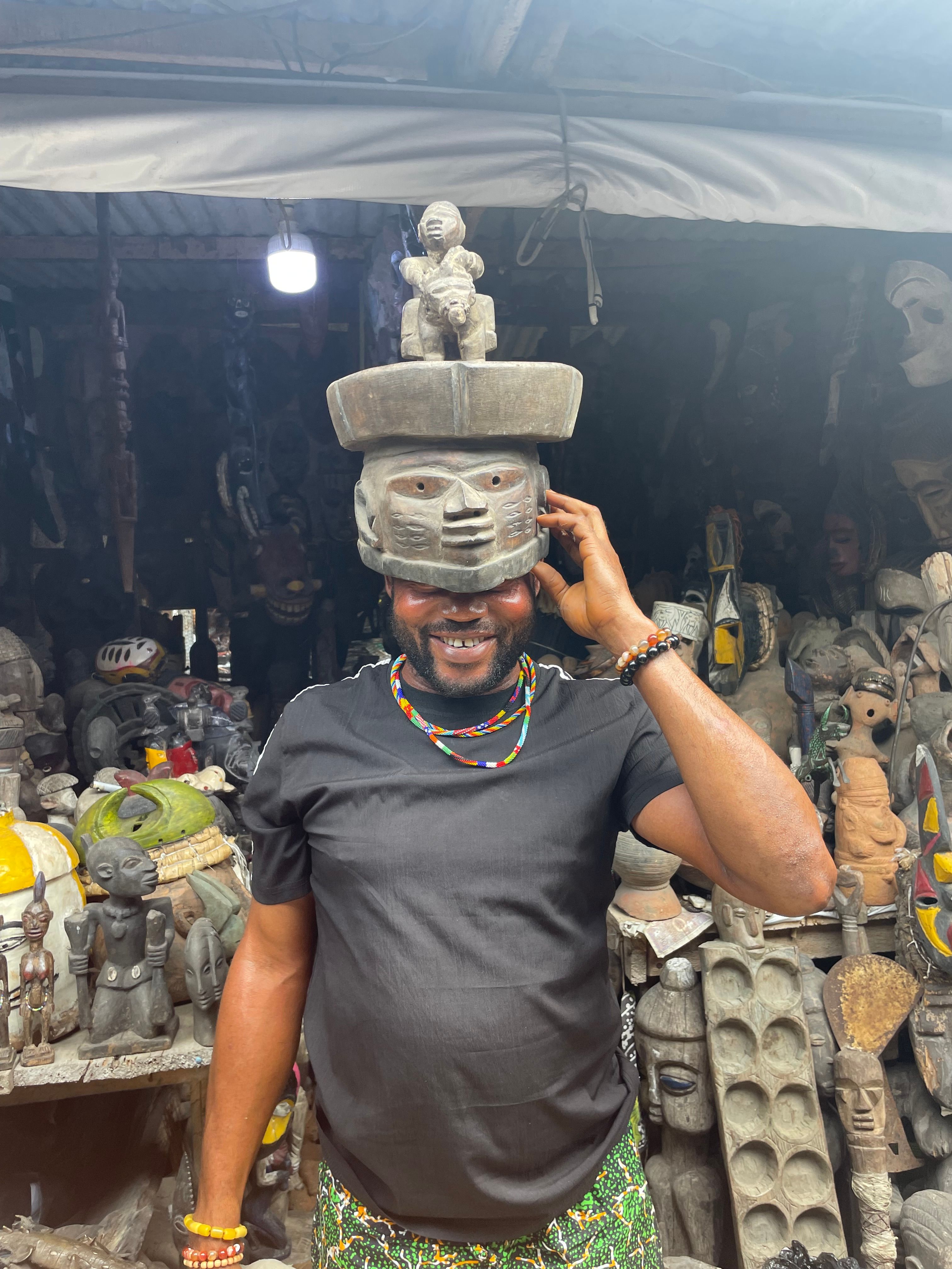 A smiling man wearing a traditional carved wooden mask in a market filled with sculptures.