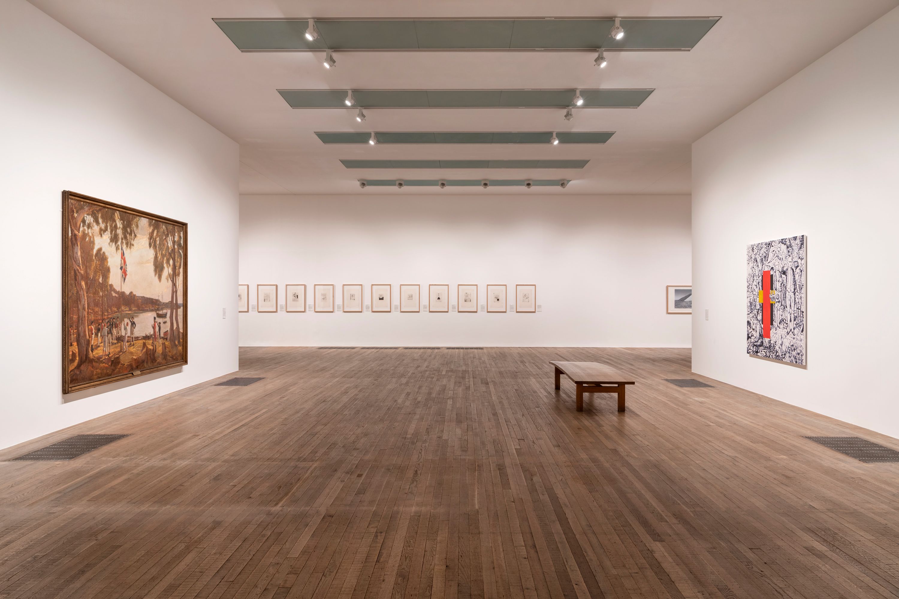 A solemn wooden bench rests in an art gallery with polished wood flooring, adorned by artwork on white walls.