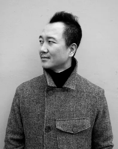 A black and white portrait of a person with short dark hair wearing a houndstooth coat, looking to the side against a plai...