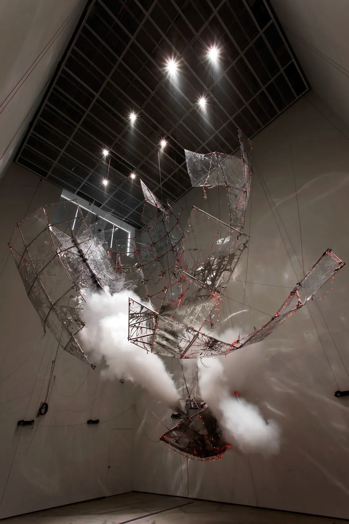 An art installation depicts transparent pieces resembling rocket parts hanging from a ceiling in a dimmed room. Smoke can be seen emanating from the grey nose cone of a rocket.