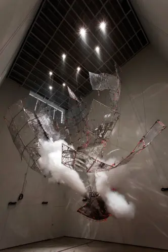 An art installation depicts transparent pieces resembling rocket parts hanging from a ceiling in a dimmed room. Smoke can ...