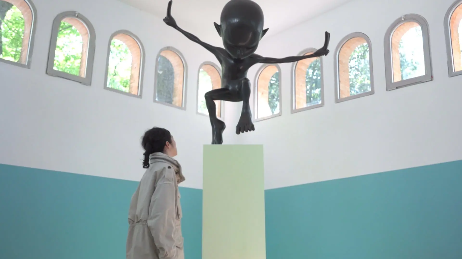 A person in a light jacket gazes at a large black sculpture of a humanoid figure in a dynamic pose on a pedestal.
