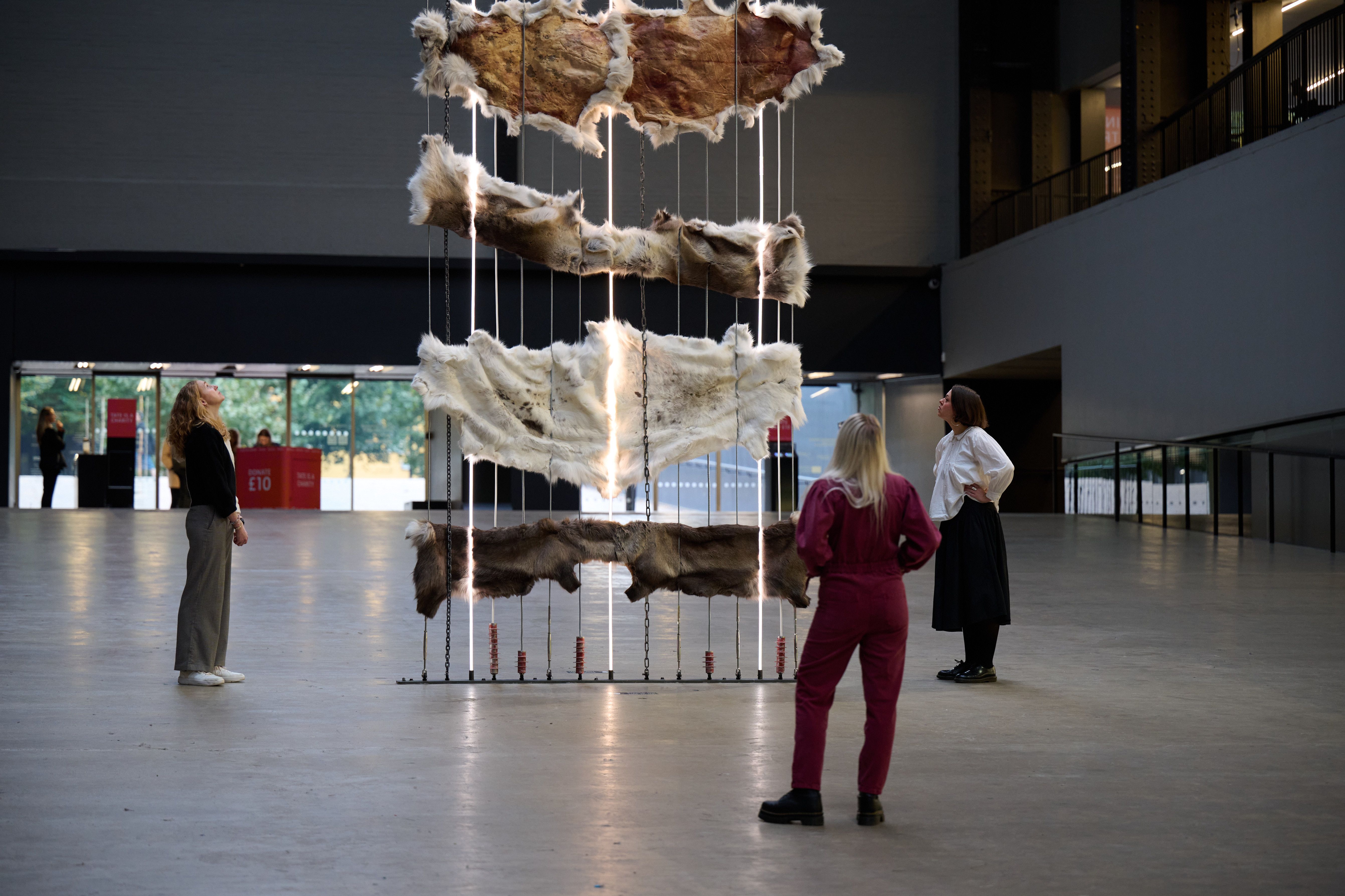 Three visitors stand around a suspended arrangement of animal hides and lights, observing it in a spacious museum atrium.