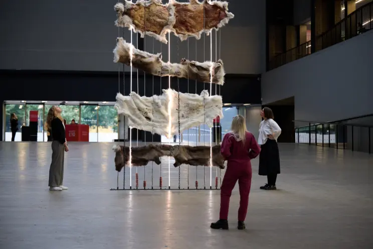 Three visitors stand around a suspended arrangement of animal hides and lights, observing it in a spacious museum atrium.