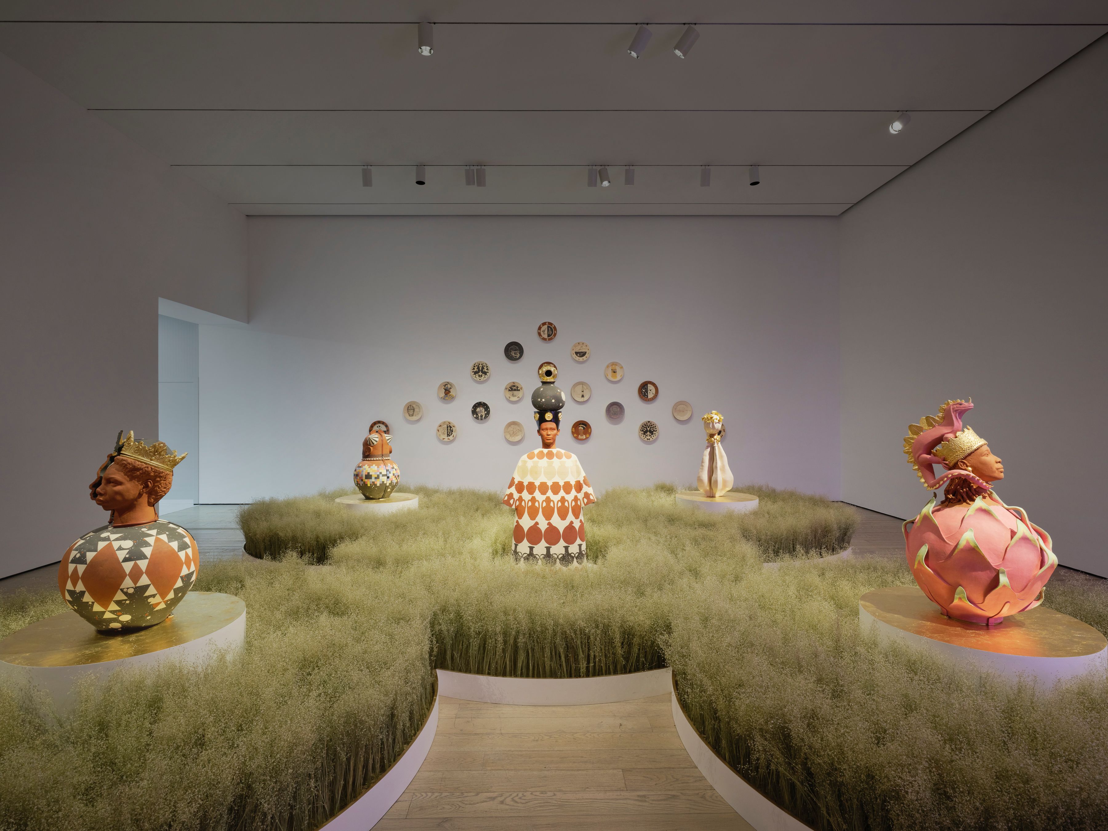 Gallery with crowned ceramic busts on patterned vessels set among soft grass mounds, with decorative plates on back wall.”