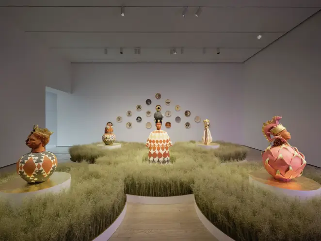 Gallery with crowned ceramic busts on patterned vessels set among soft grass mounds, with decorative plates on back wall.”