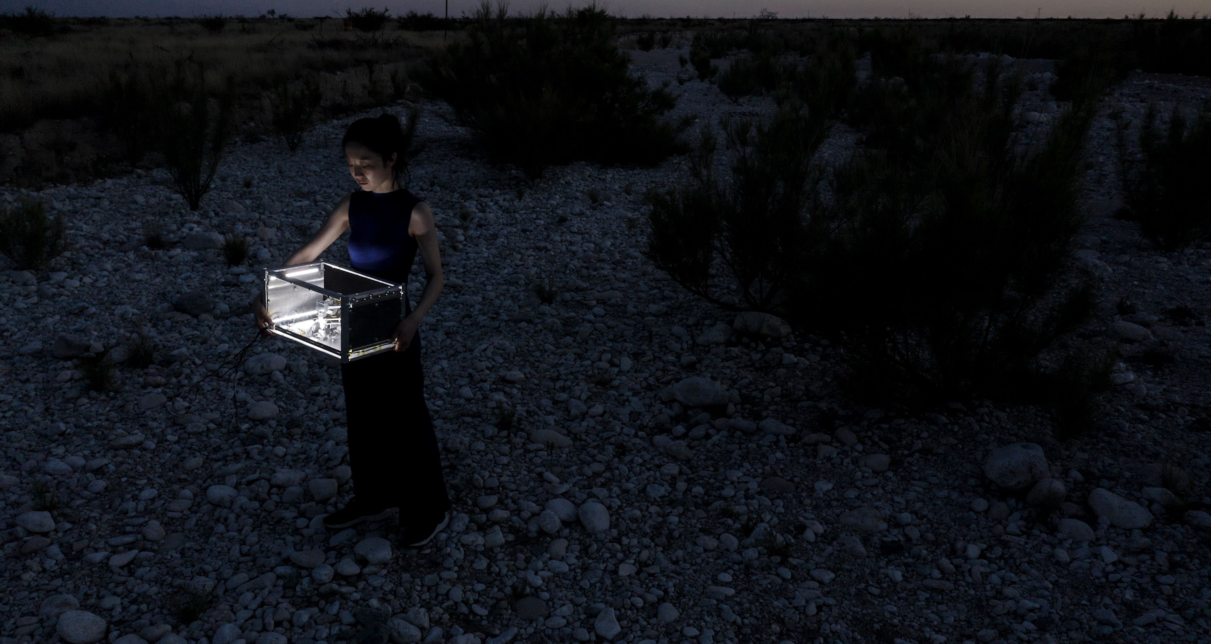 Person standing in a rocky desert at night holding an illuminated transparent box.