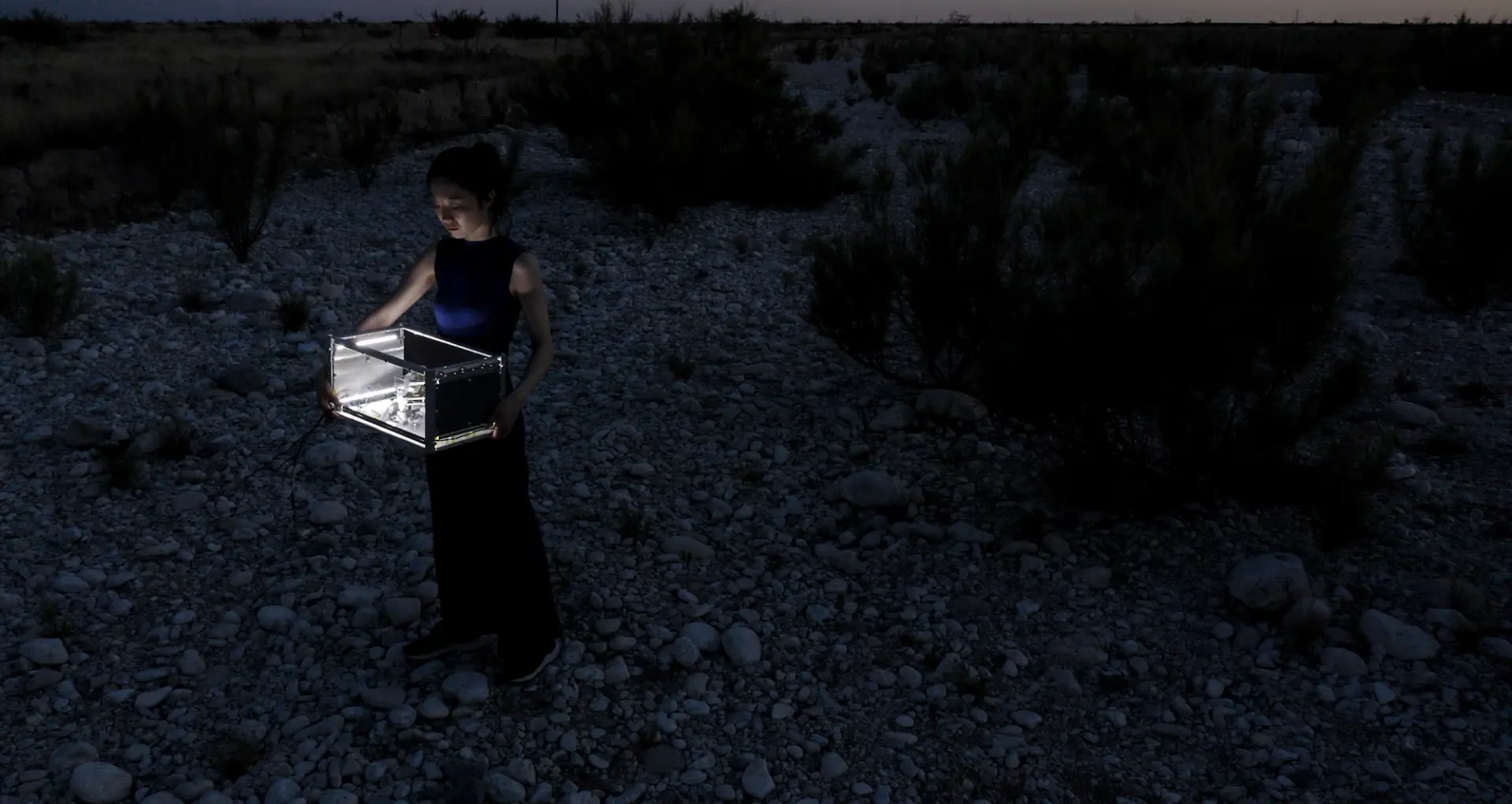Person standing in a rocky desert at night holding an illuminated transparent box.