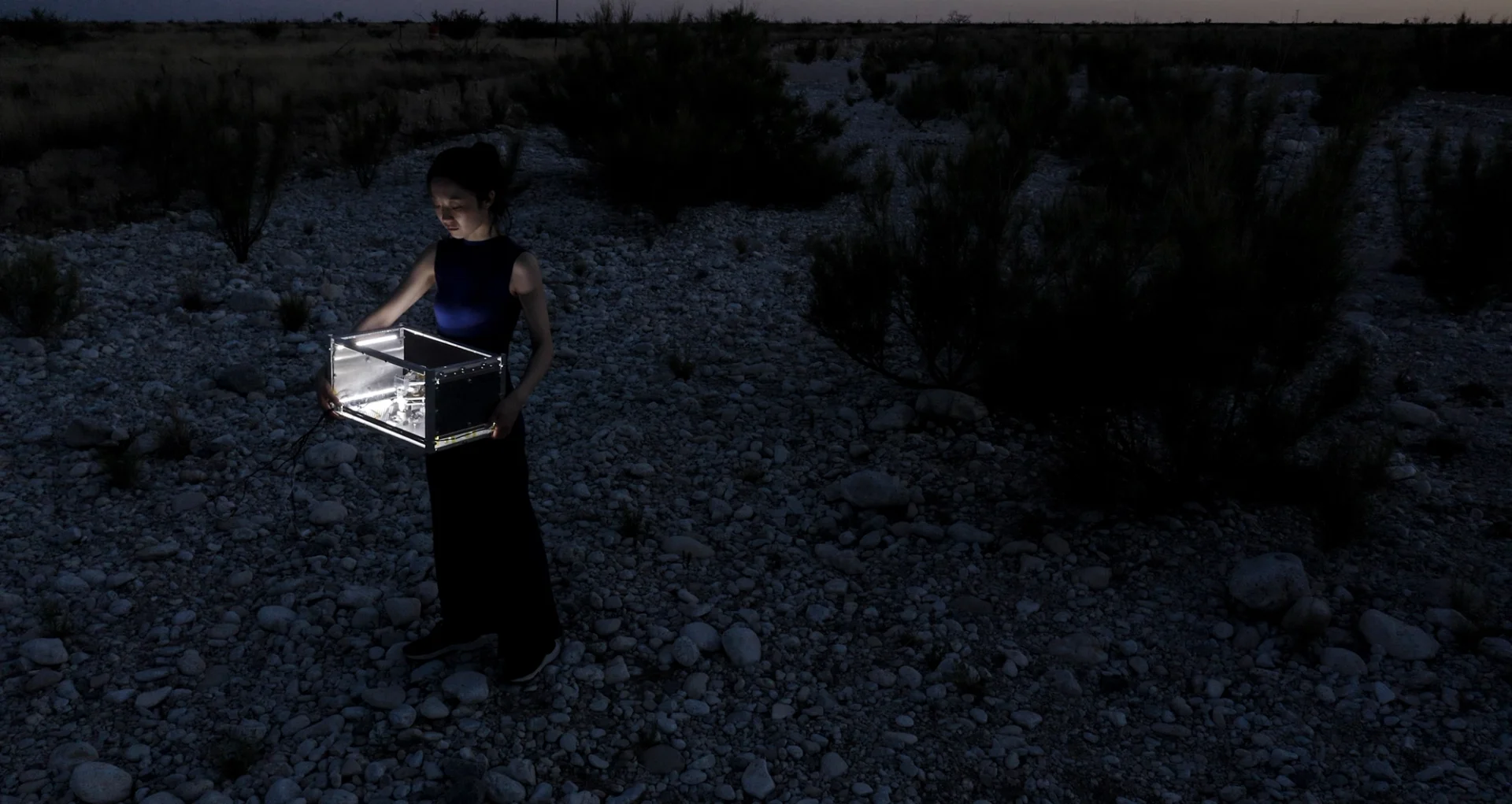 Person standing in a rocky desert at night holding an illuminated transparent box.
