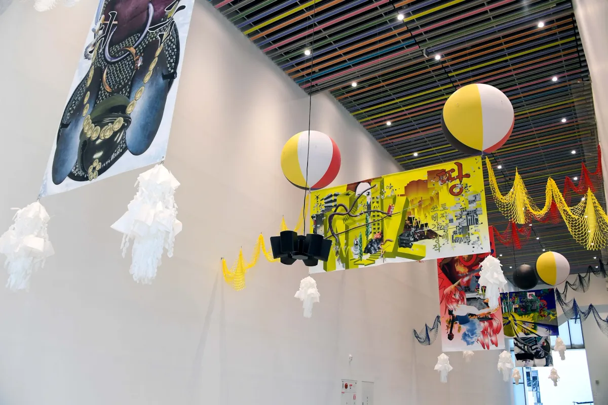 Art installation suspended from a ceiling featuring nets in various colors, white ornaments, and two multi-colored balls. The scene includes graphic colorful posters.