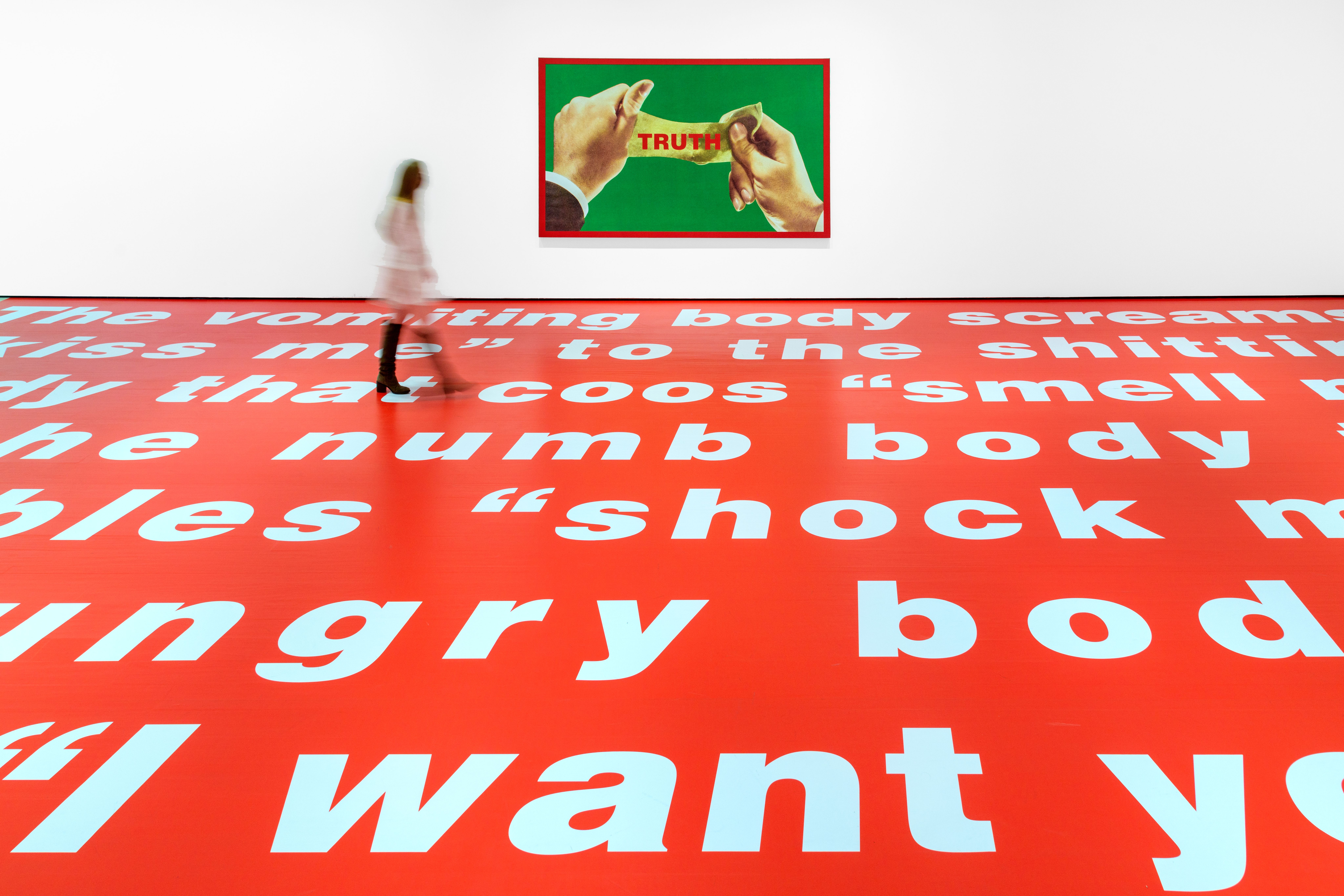 A blurred human figure strides across a vibrant red floor embellished with striking white text. Behind them, prominently d...