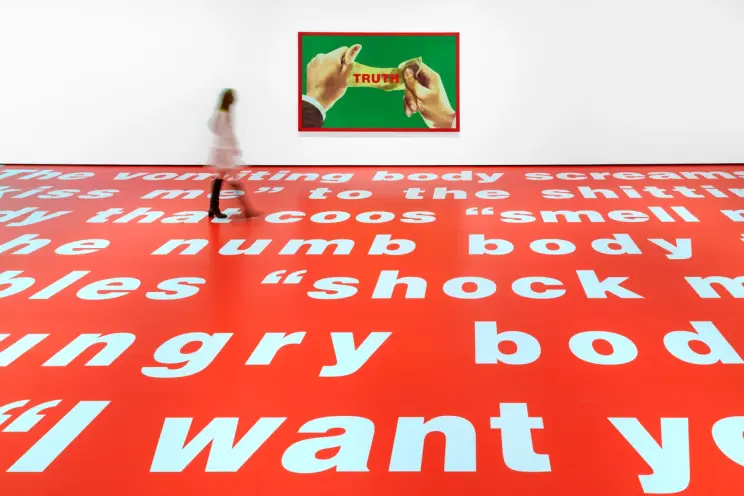 A blurred human figure strides across a vibrant red floor embellished with striking white text. Behind them, prominently d...