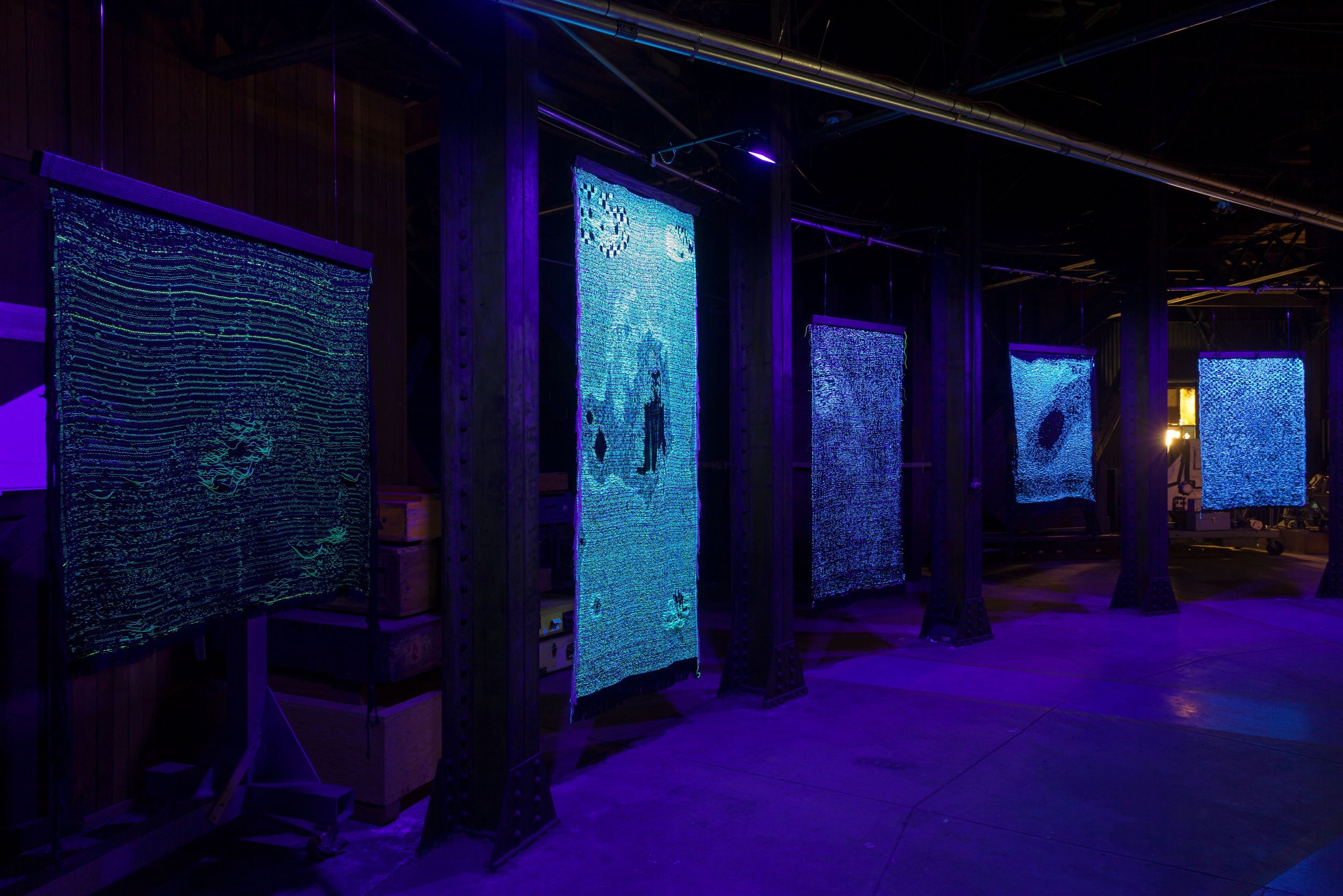 A series of textile artworks illuminated under blue lights, suspended in an industrial exhibition space.