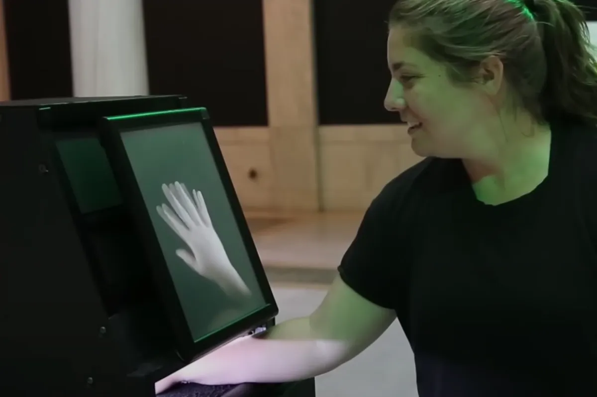 A person places their hand into a machine incorporating a video screen, which displays an image of a hand.