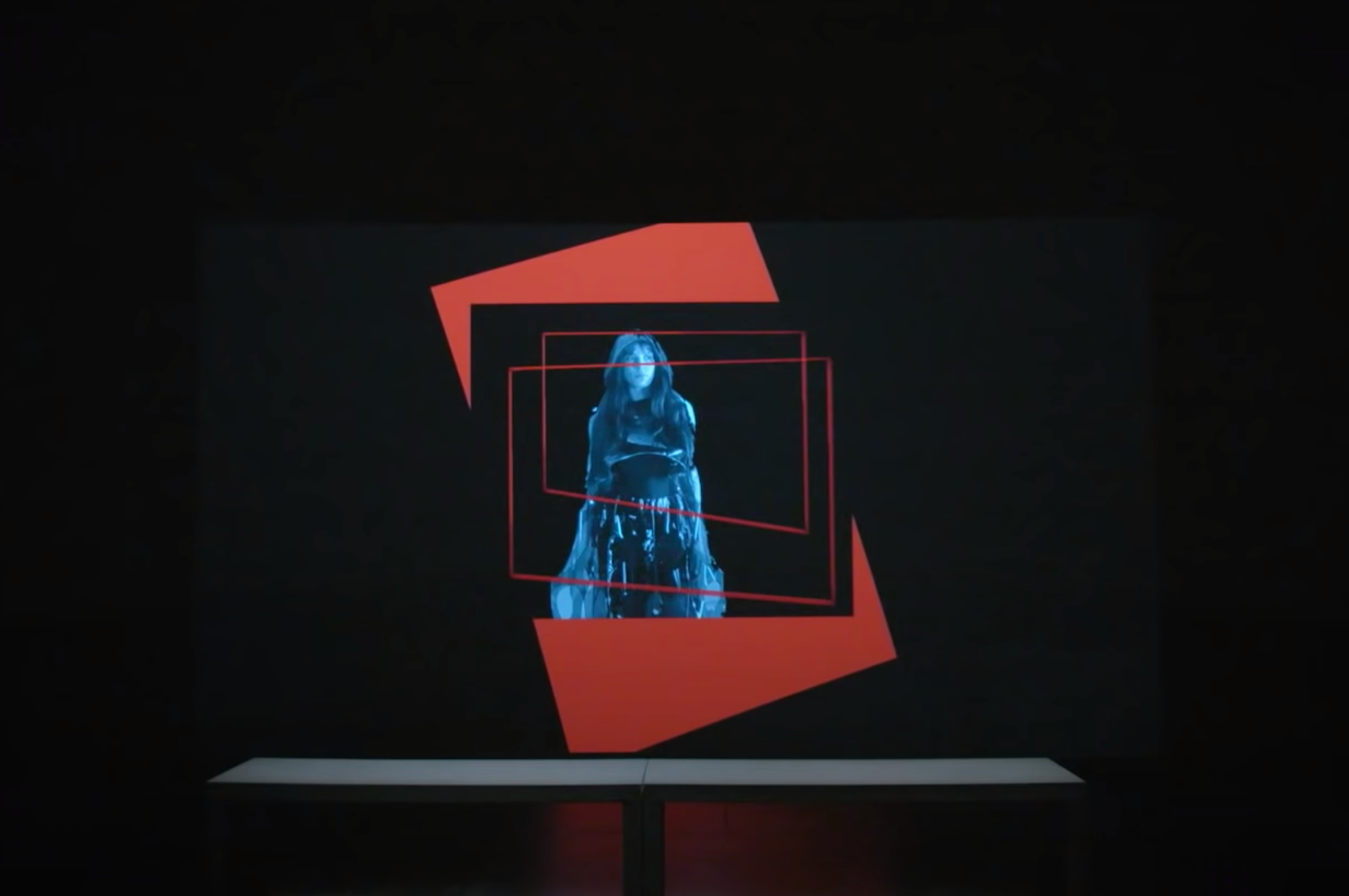 A dark room features a projected art piece with multiple layers: a red vertical rectangle leans to the left against a blac...