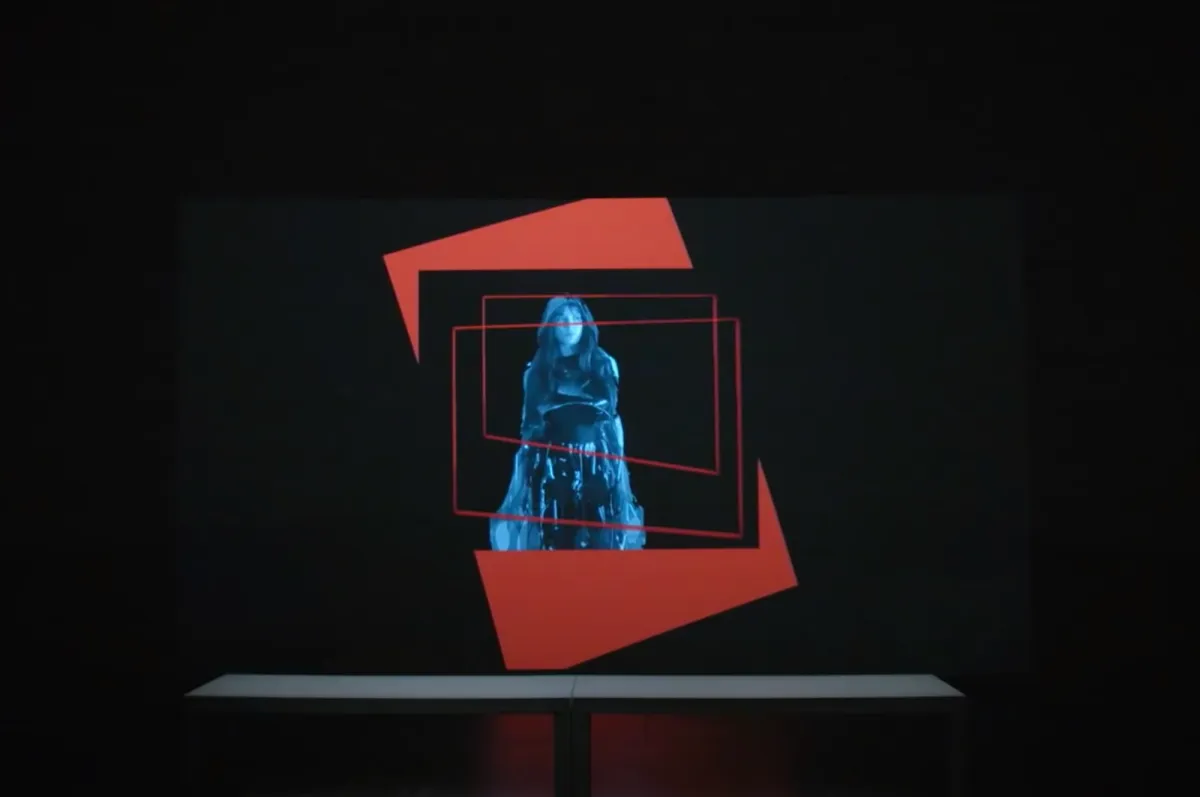 A dark room features a projected art piece with multiple layers: a red vertical rectangle leans to the left against a black backdrop, overlaid with a smaller black square holding a cut-out, blue-tinted image of a person. The top layer consists of two irregularly shaped red rectangle borders.