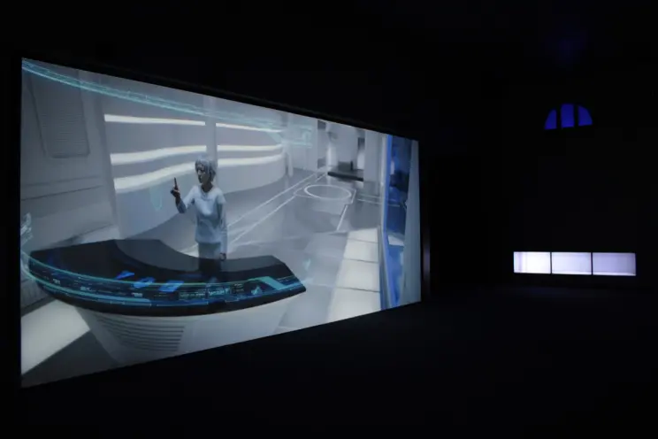 A projected image of an individual dressed in white, situated in a well-lit laboratory environment, holds their index fing...