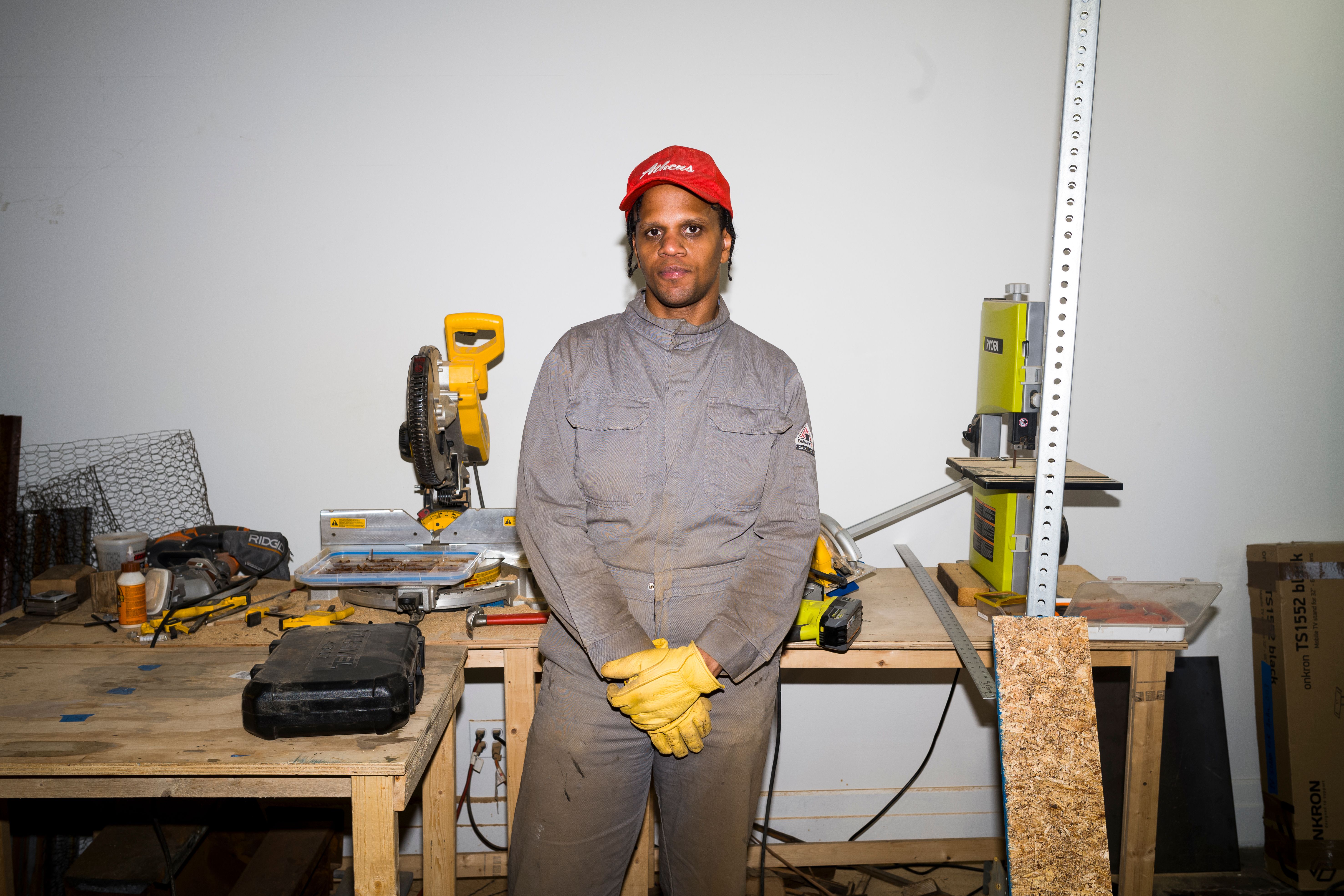 Person in gray jumpsuit and red hat standing in a workshop with tools in the background.