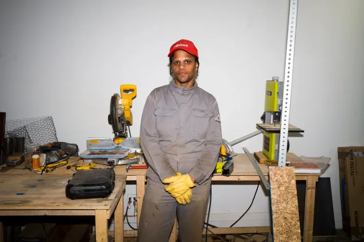 Person in gray jumpsuit and red hat standing in a workshop with tools in the background.