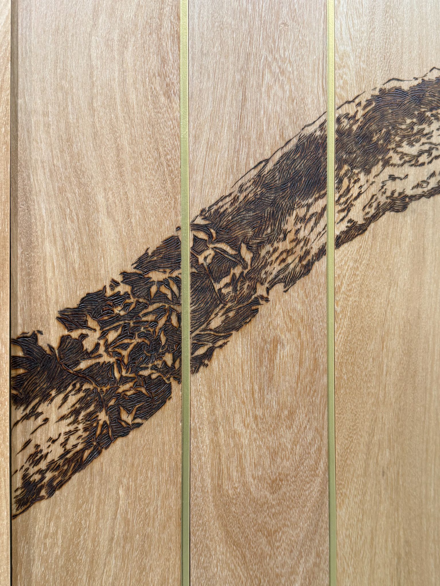 Close-up of a pyrographed (burnt) tree branch pattern on smooth wood panels with brass lines.