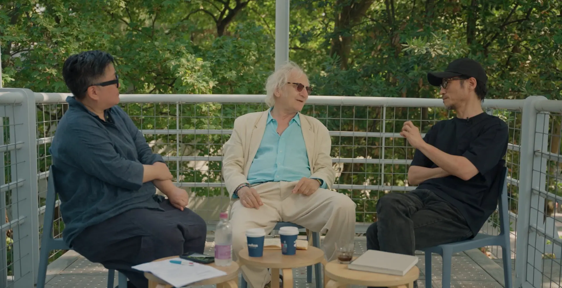 Three men engaged in conversation, seated outdoors on a balcony surrounded by lush greenery.