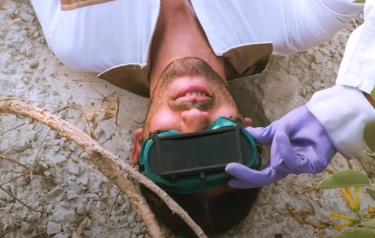 A person in white clothing lies on white soil scattered with twigs and fallen leaves, smiling as they hold a green eye protector on their eyes. Their hand is covered with a lilac rubber glove.