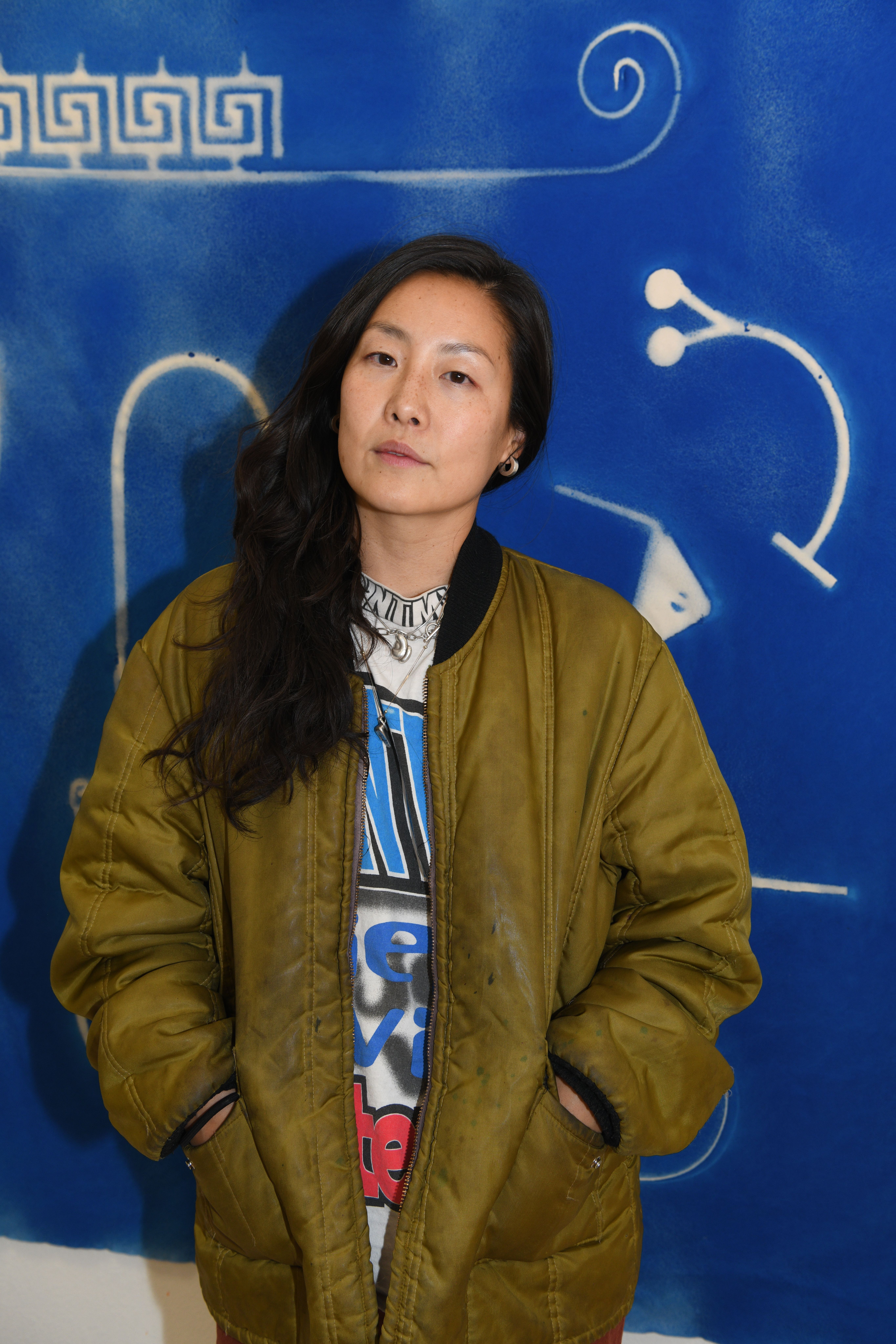 Artist standing against a vibrant blue backdrop with abstract glyphs, conveying a strong, contemplative presence.
