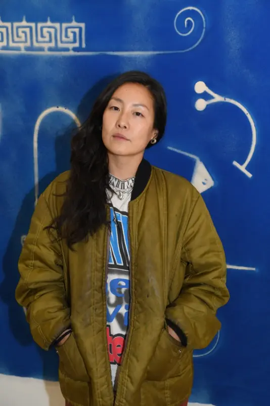 Artist standing against a vibrant blue backdrop with abstract glyphs, conveying a strong, contemplative presence.