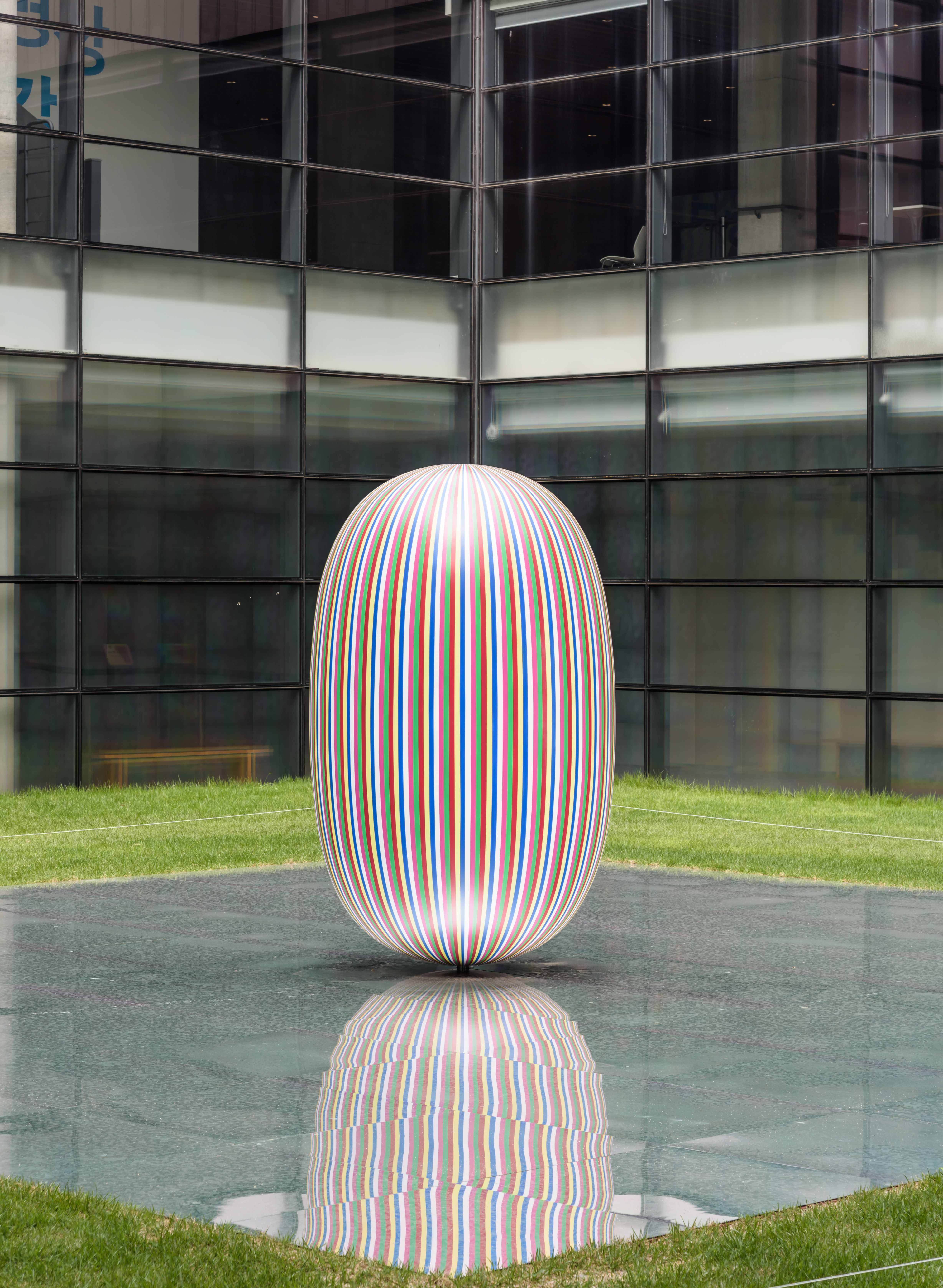 An elliptical sculpture is displayed on a mirrored rectangular surface in a building's backyard, surrounded by many windows.