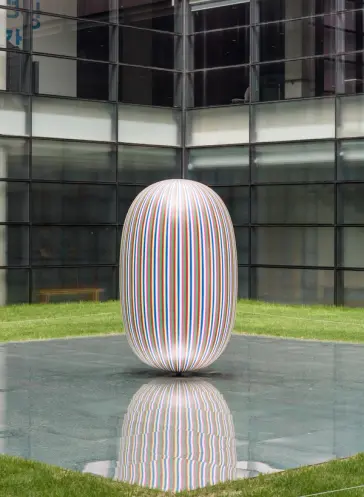An elliptical sculpture is displayed on a mirrored rectangular surface in a building's backyard, surrounded by many windows.