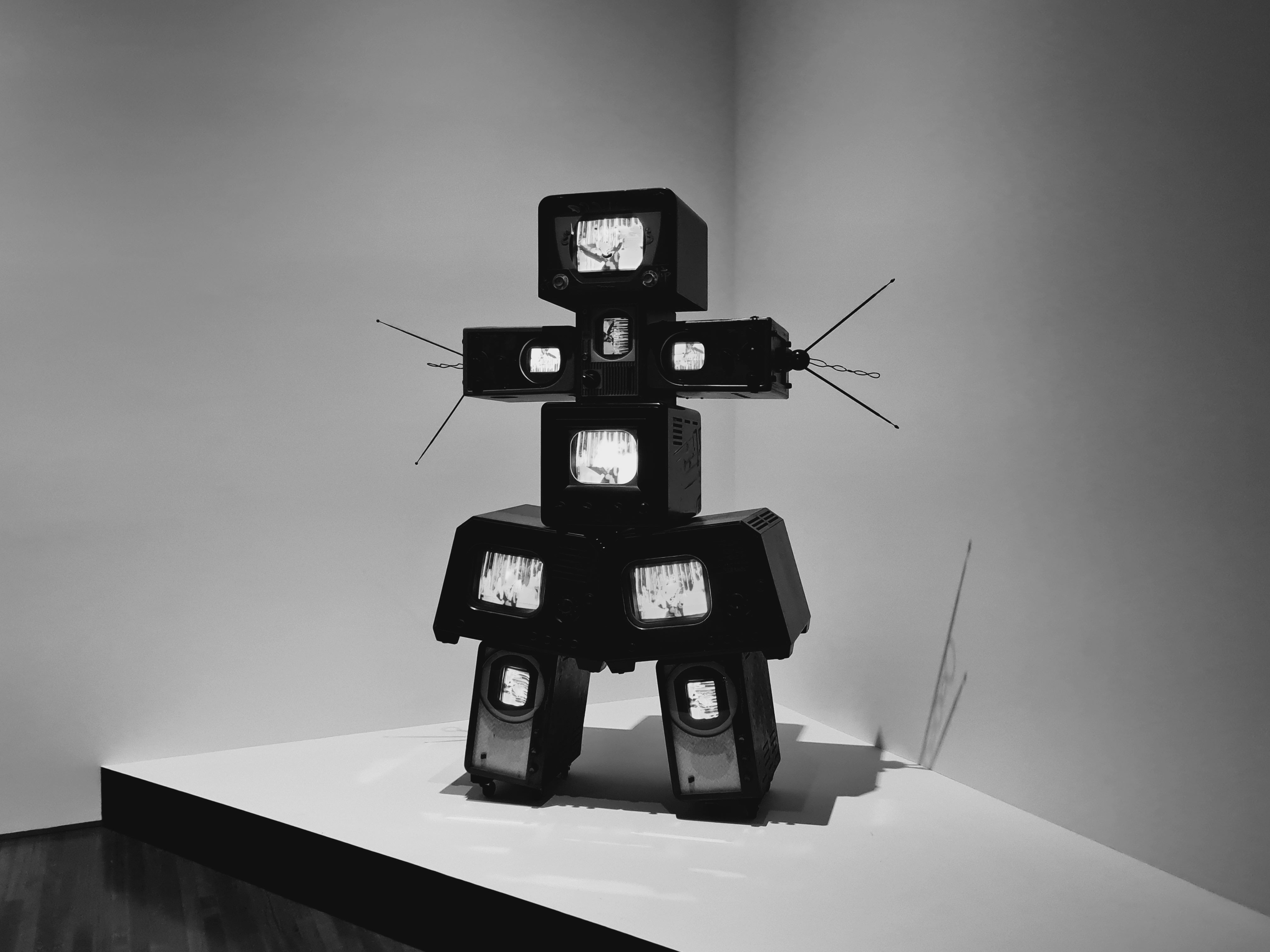 A modern art sculpture made of stacked retro televisions displaying static, arranged to resemble a humanoid figure, displayed in a gallery space.