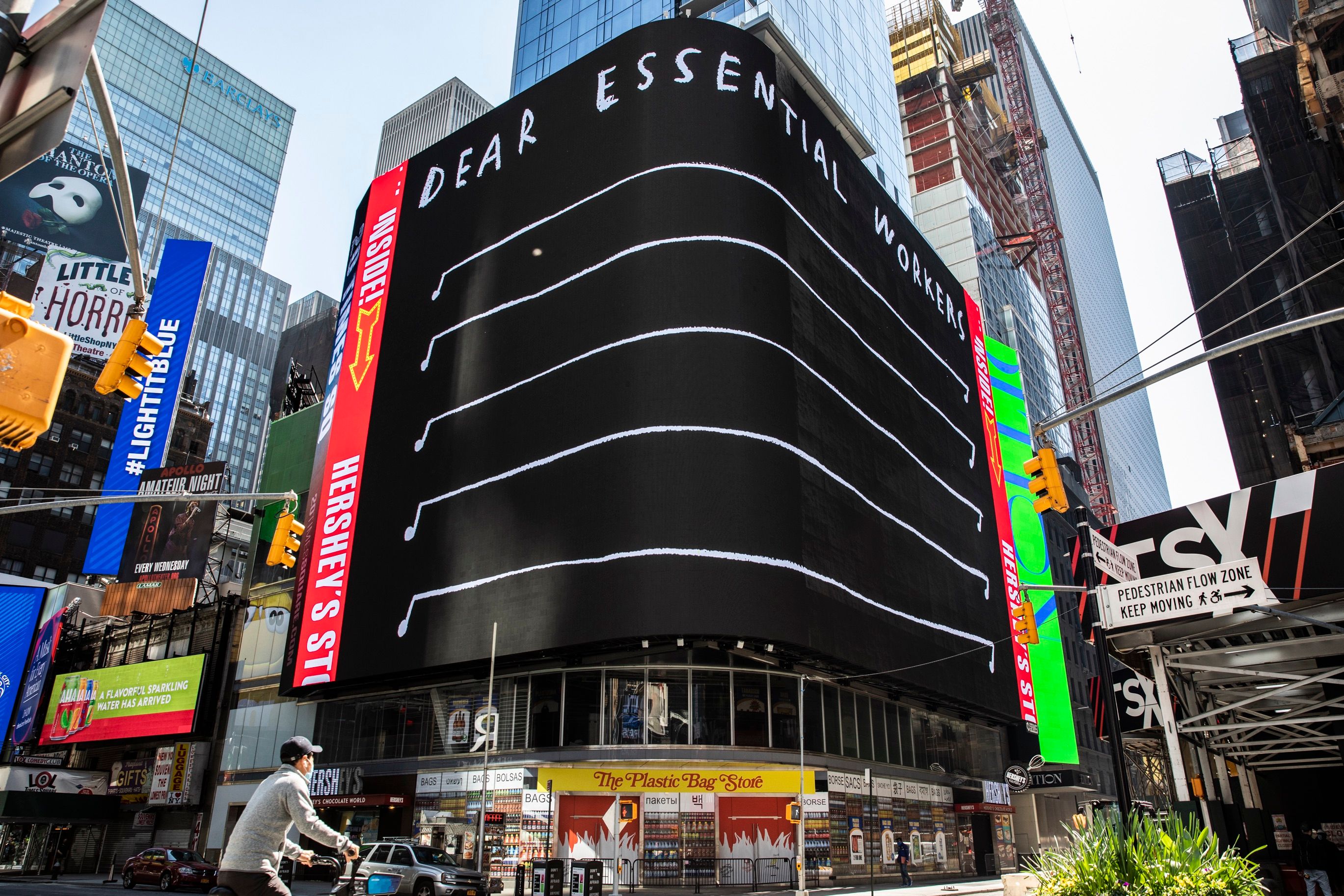 A massive digital billboard in Times Square reads “DEAR ESSENTIAL WORKERS” above a series of musical staff-like lines.