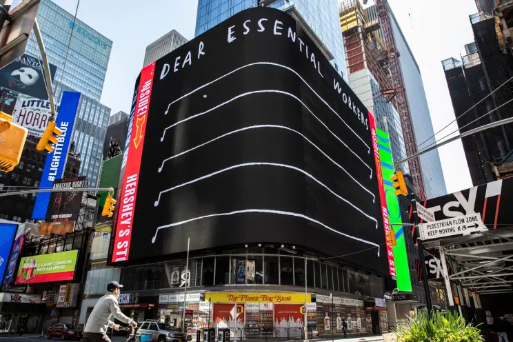 A massive digital billboard in Times Square reads “DEAR ESSENTIAL WORKERS” above a series of musical staff-like lines.