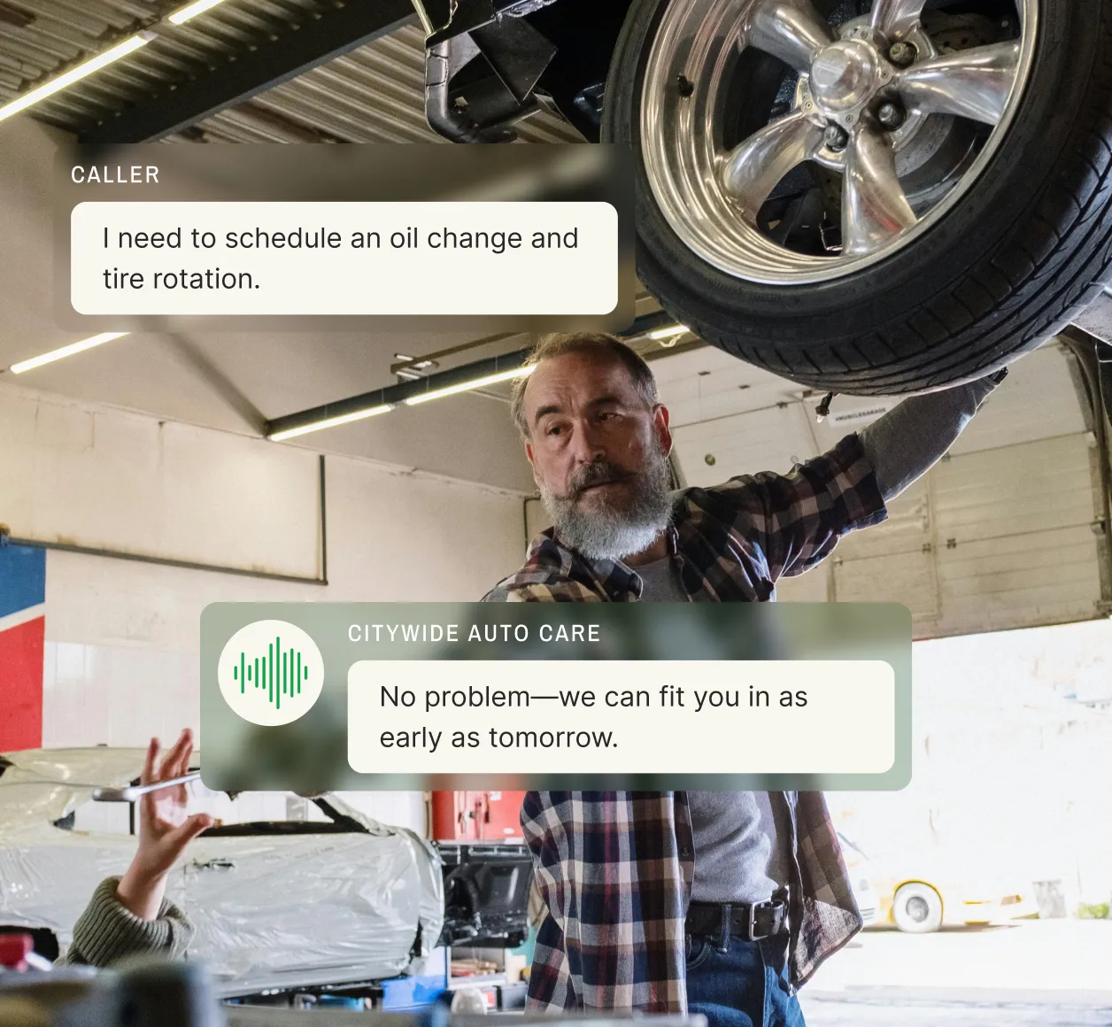 An auto mechanic works under a car in a repair shop while using voicemail transcription to confirm a voice message about scheduling an oil change and tire rotation.