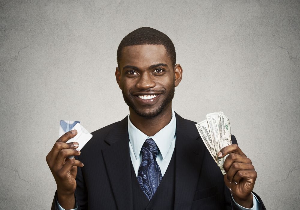 Closeup portrait happy%2c smiling business man company employee standing%2c holding dollar bills%2c credit card hand isolated grey black background. Banking exchange rate concept. Facial expression reaction.jpeg